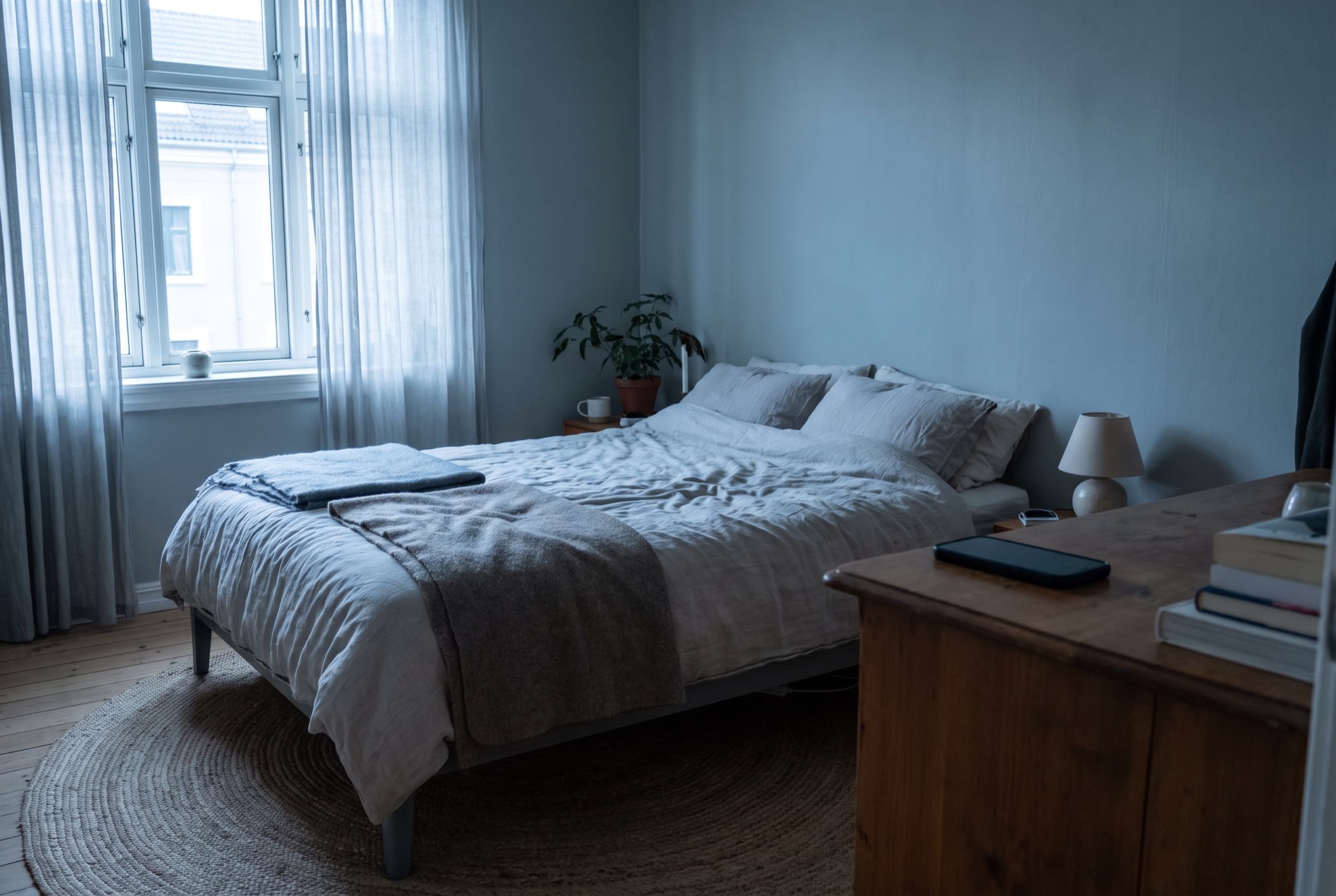 A dark bedroom at blue hour, cool light grazing rumpled linen and a wooden sill