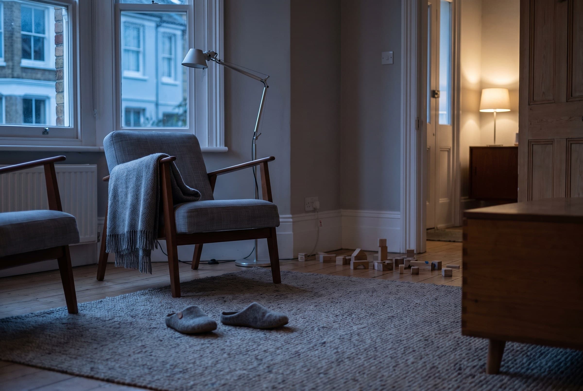 A living room after bedtime with grey slippers kicked off on a woven rug, a wool throw over an empty armchair, plain wooden blocks scattered by the skirting and one lamp lit in the next room