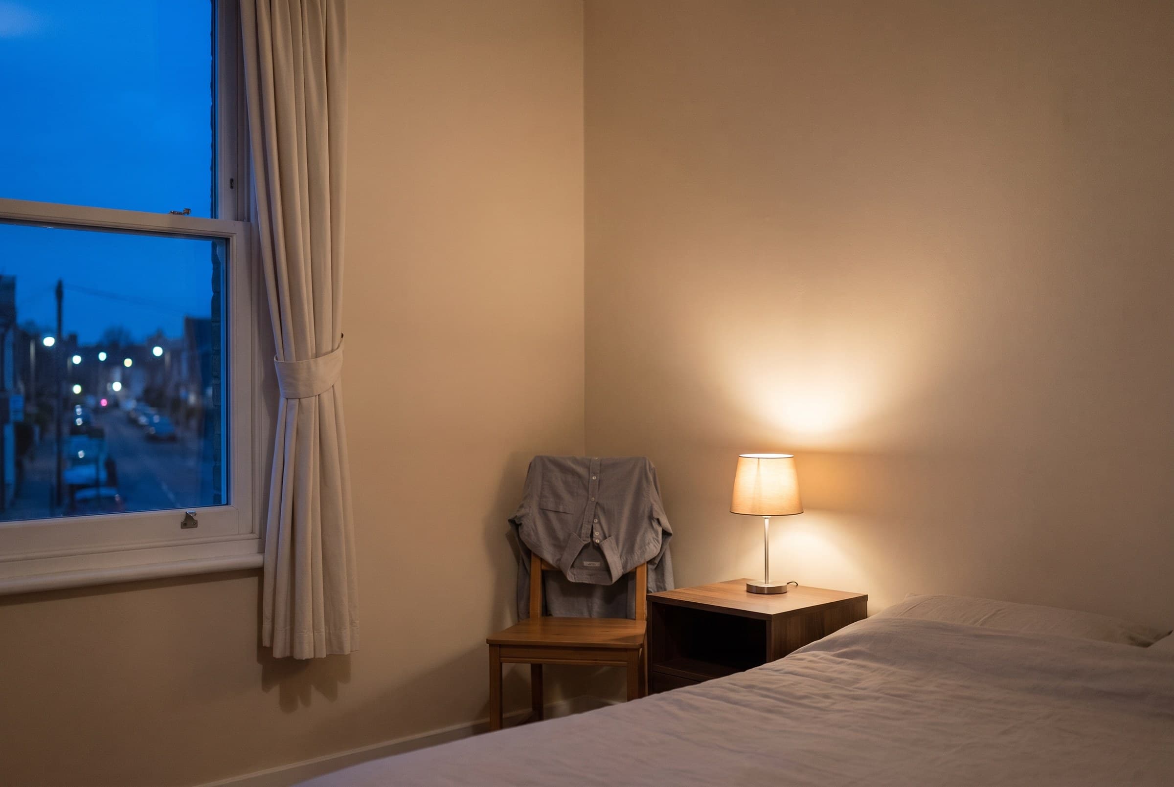 A tidy bedroom at dusk with the curtain tied back over a blue street, one small shaded lamp burning low and a shirt laid over a wooden chair