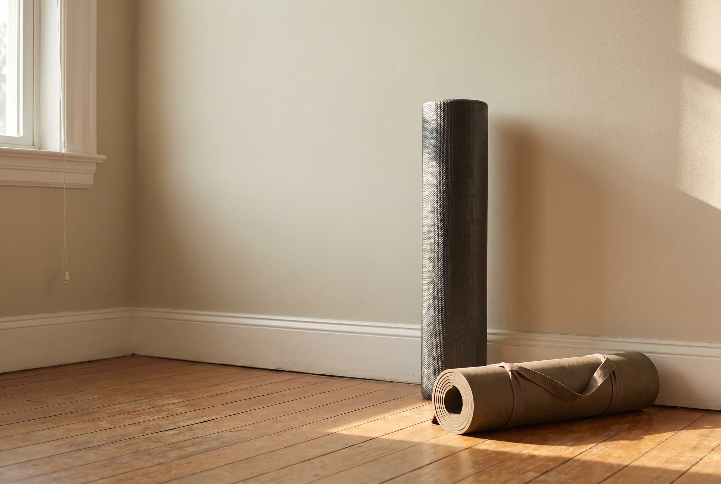 A foam roller standing on end against a pale wall beside a rolled exercise mat, morning light grazing the foam across a wooden floor