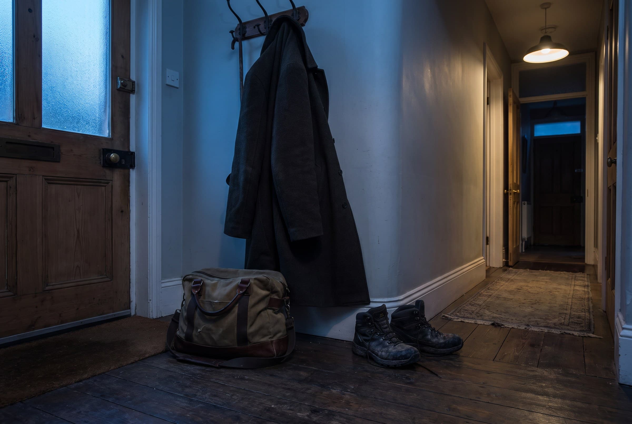 A dim hallway entry at dusk with a canvas work bag dropped on bare boards, a heavy coat half off its hook and muddy boots kicked off beside it
