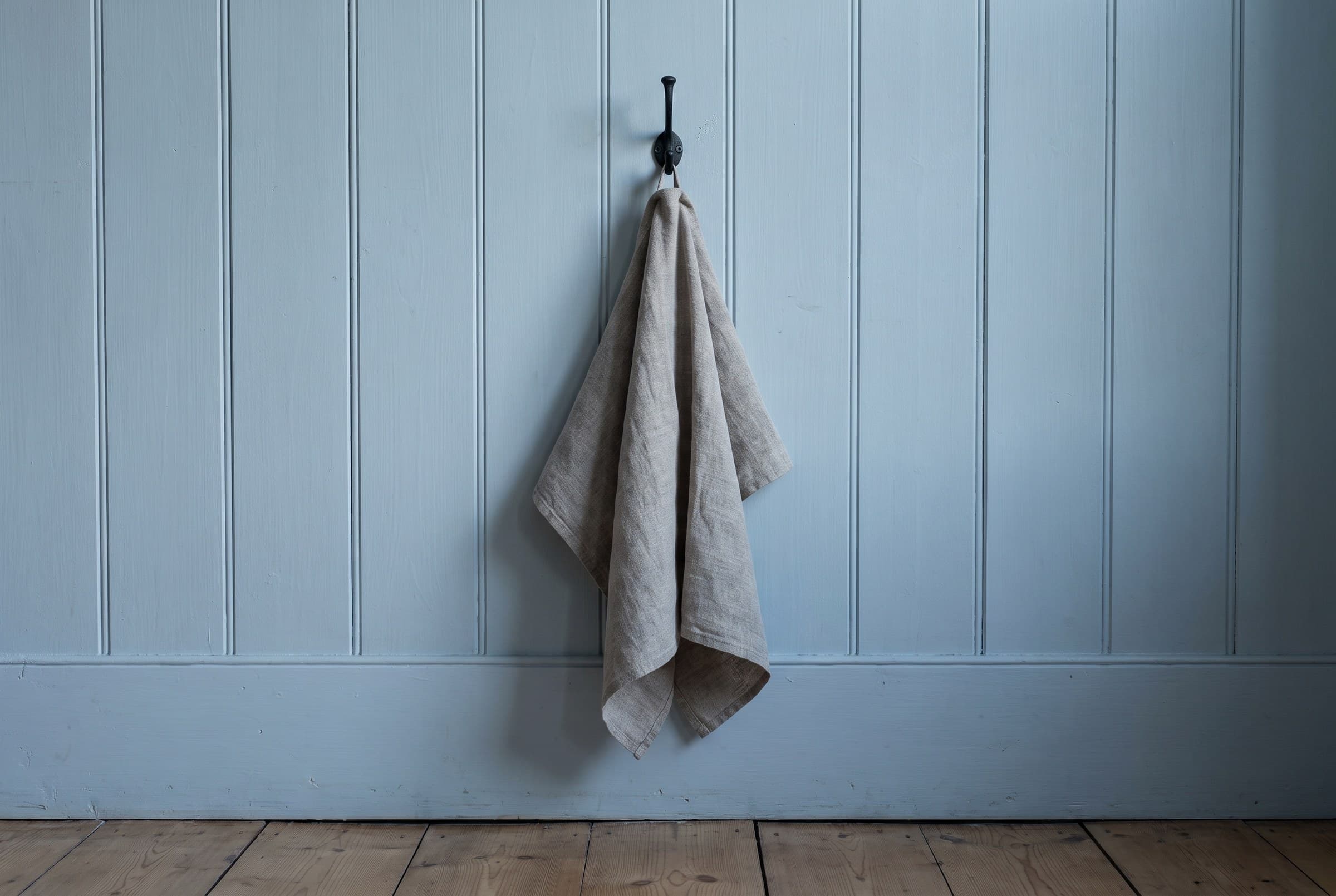 A folded towel hanging from a black iron hook on pale-blue tongue-and-groove boards above clean floorboards in cool evening light
