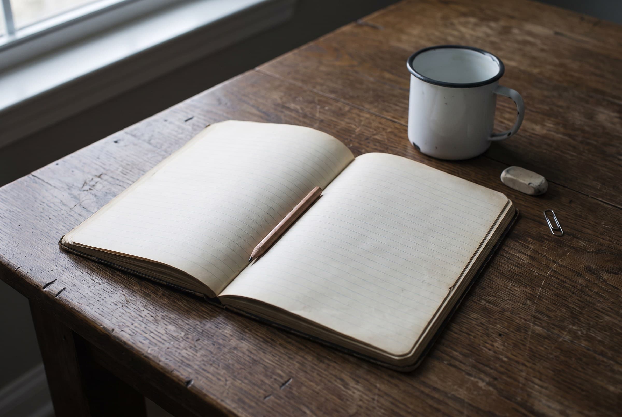 An open ruled notebook with blank pages and a pencil in its gutter on a dark scarred oak table, beside a white enamel mug, an eraser stub and a paperclip