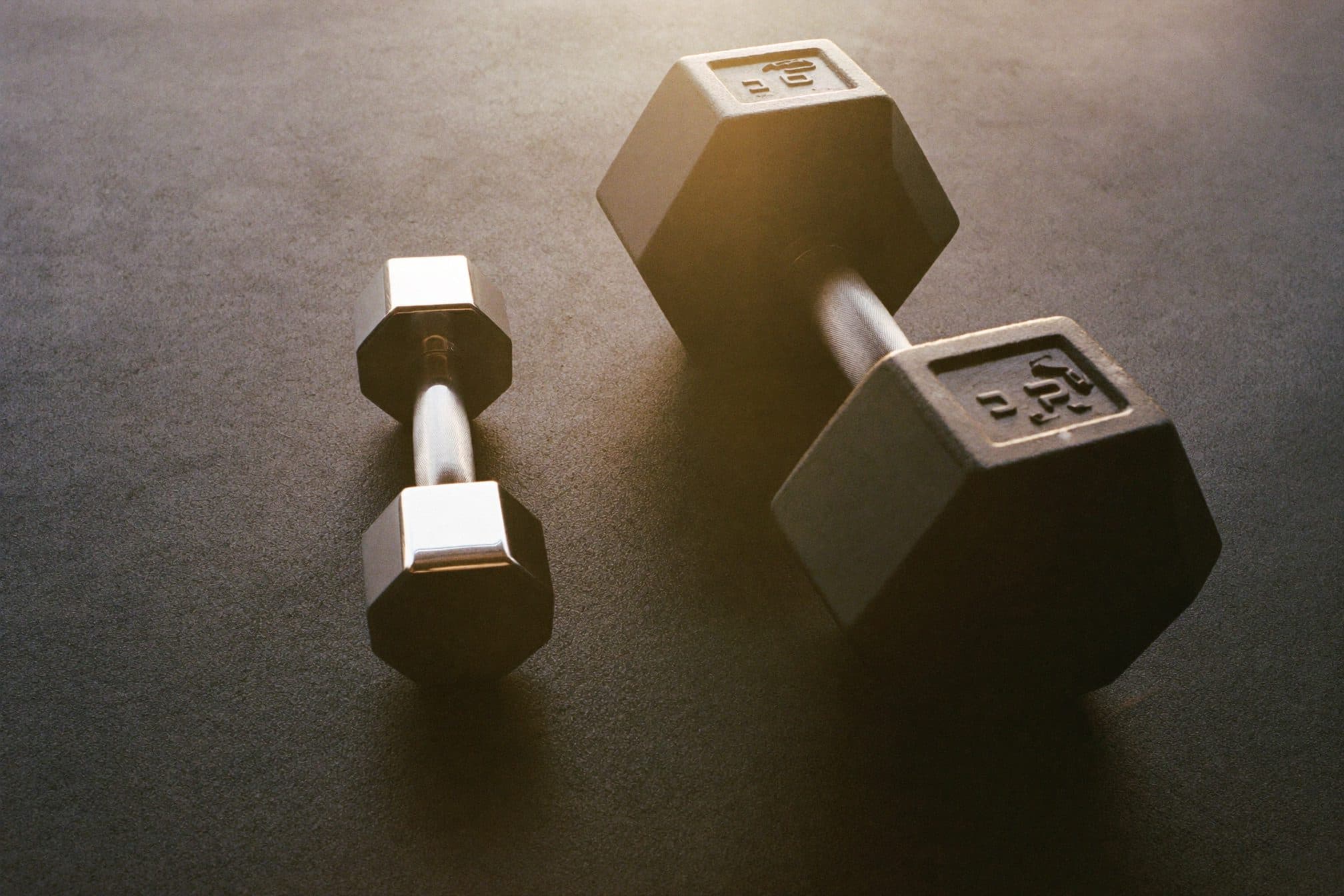 One small chrome dumbbell and one large black hex dumbbell side by side on a gym floor in morning sun
