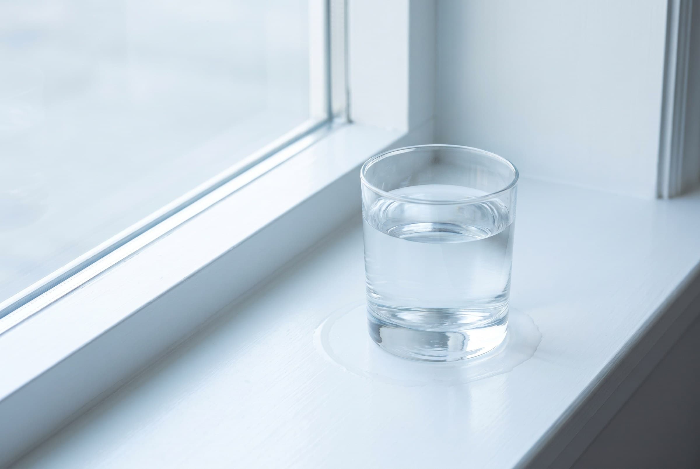 A plain glass of water standing in a faint ring of condensation on a clean white painted windowsill in cool morning light