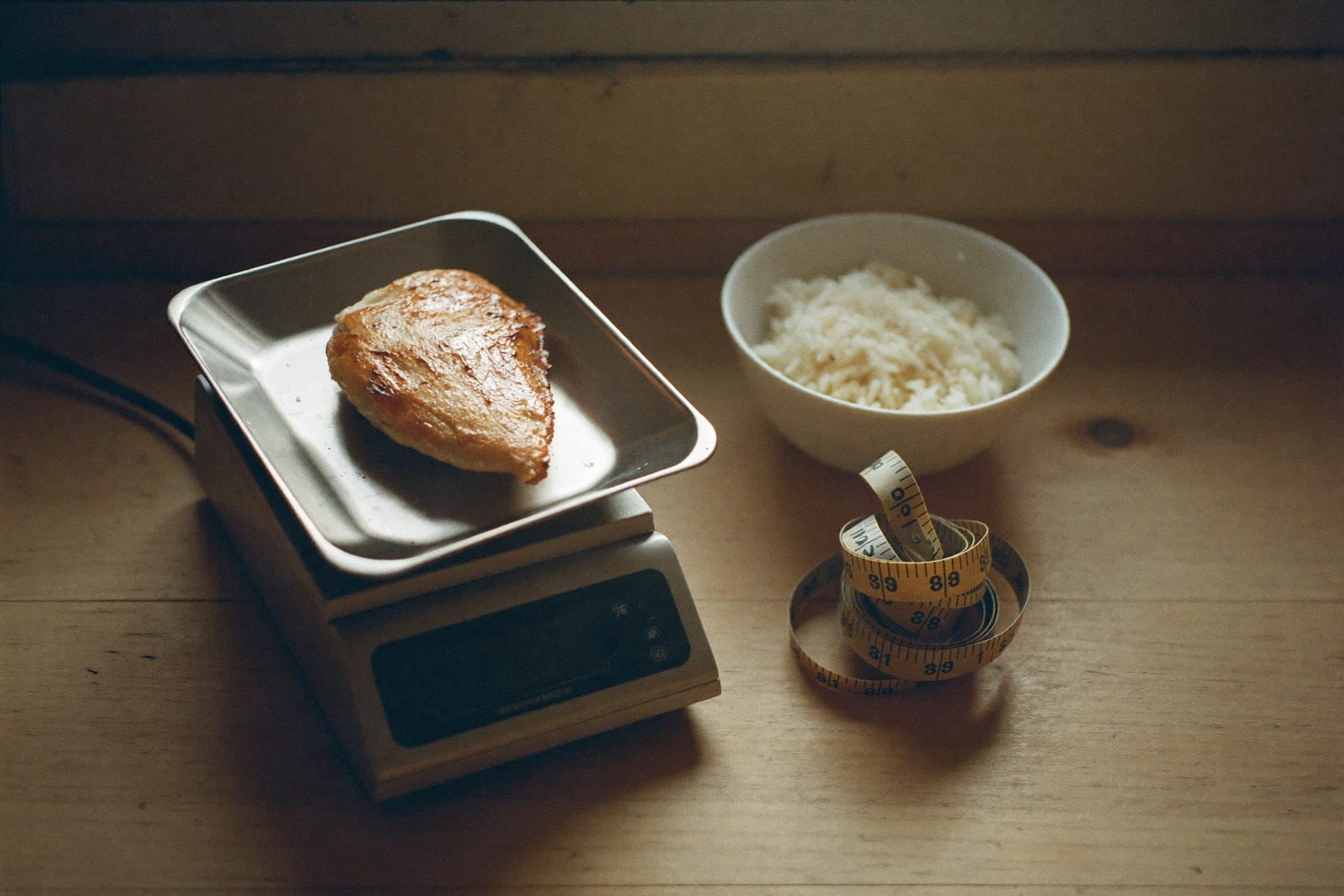 A kitchen food scale holding grilled chicken beside a small bowl of rice and a coiled tape measure in morning light