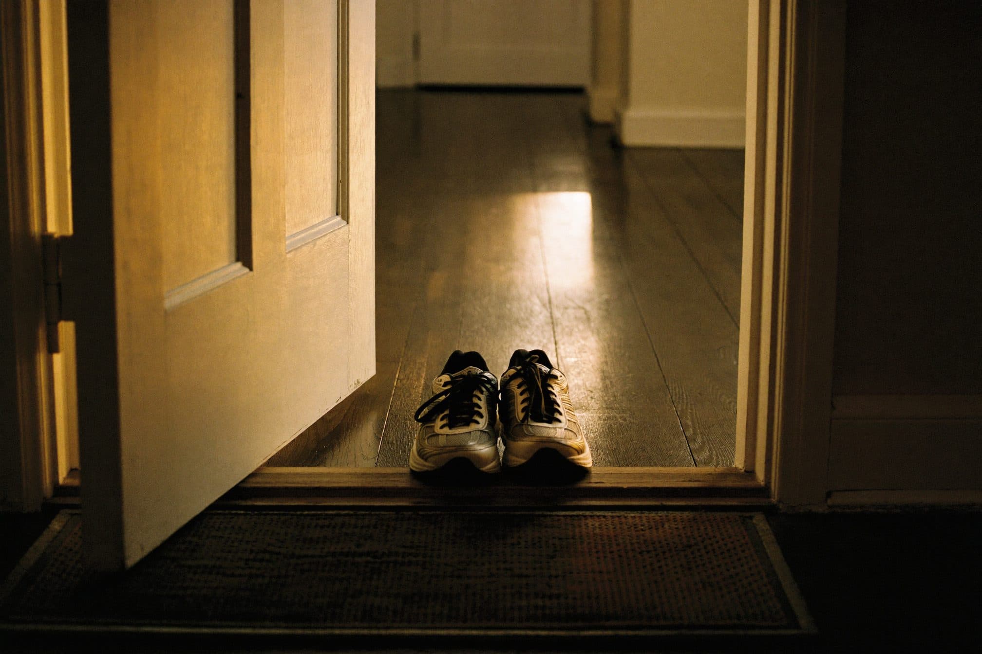 Worn running sneakers on a doormat inside an open front door, warm sunrise light across the floorboards