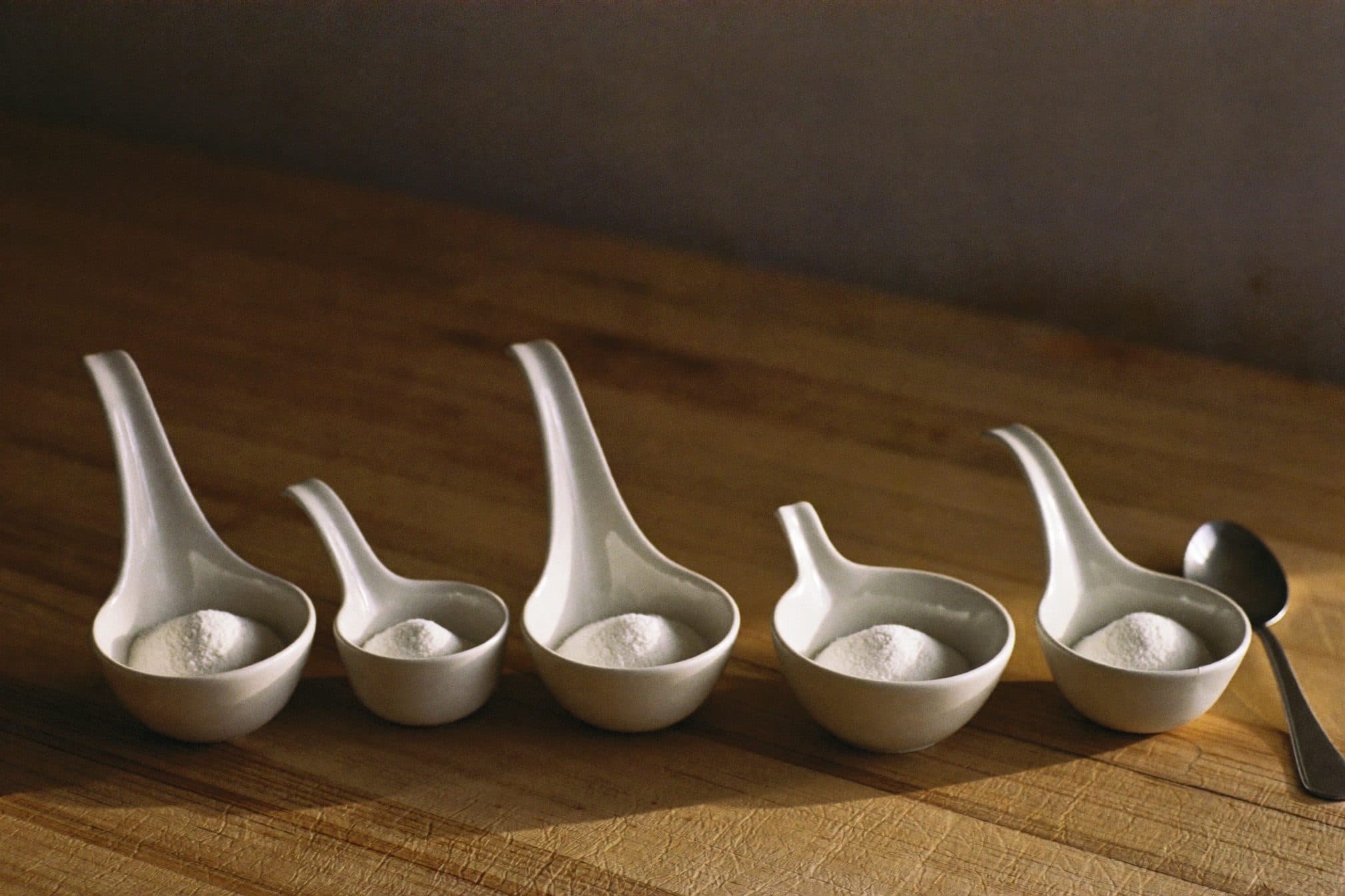 Five small white ceramic bowls in a row, each holding the same small mound of white powder, morning light