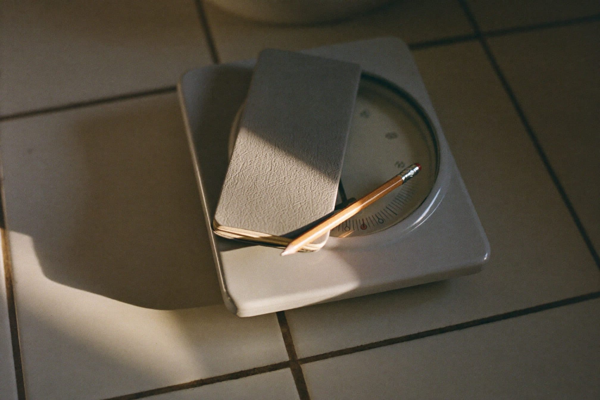 A pencil and closed pocket notebook resting on a plain bathroom scale on tile in morning light