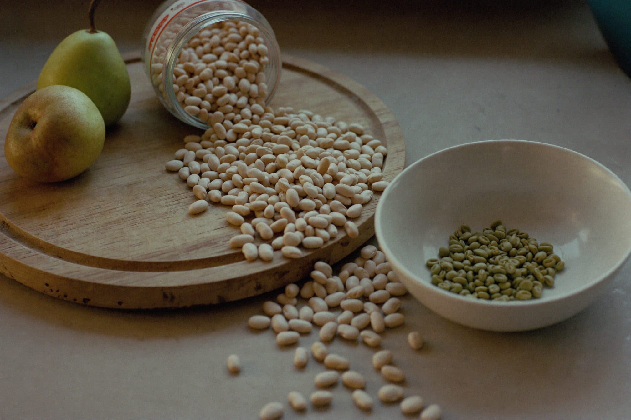 A tipped glass jar spilling pale beans across a wooden board, with a bowl of green peas and two pears