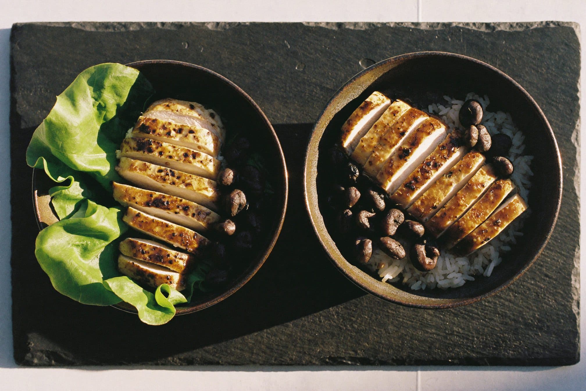 Overhead view of two dark ceramic bowls with the same grilled chicken and beans, one on lettuce and one on rice