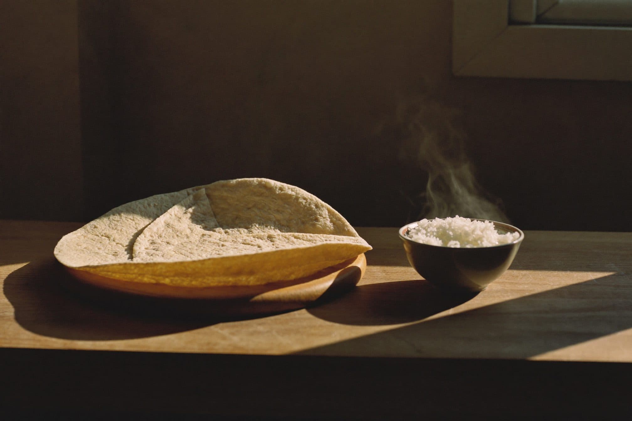A large flour tortilla draped over a wooden board beside a small bowl of steaming rice