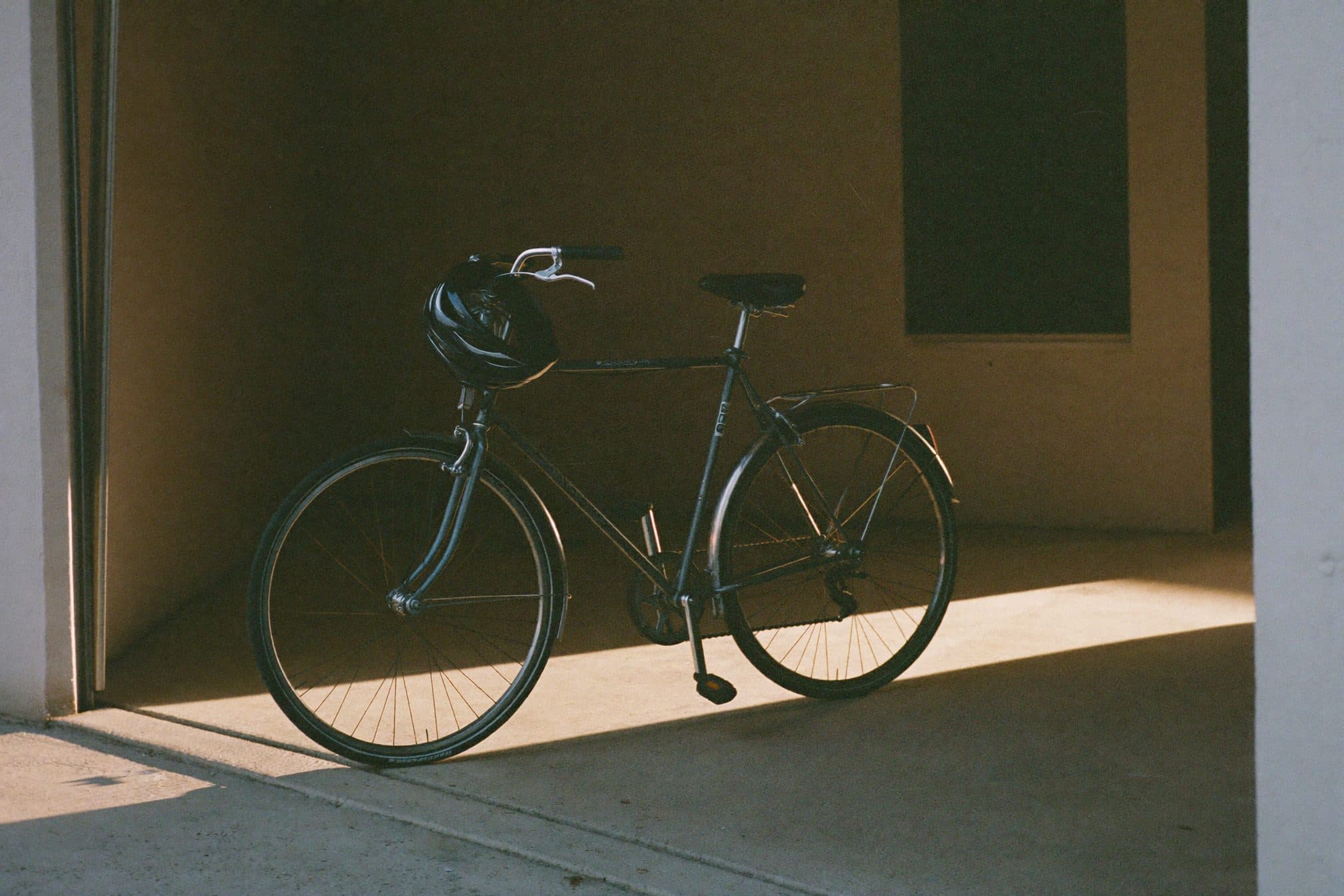 A commuter bicycle leaning in a garage doorway with a helmet on the handlebar, dawn light on the concrete