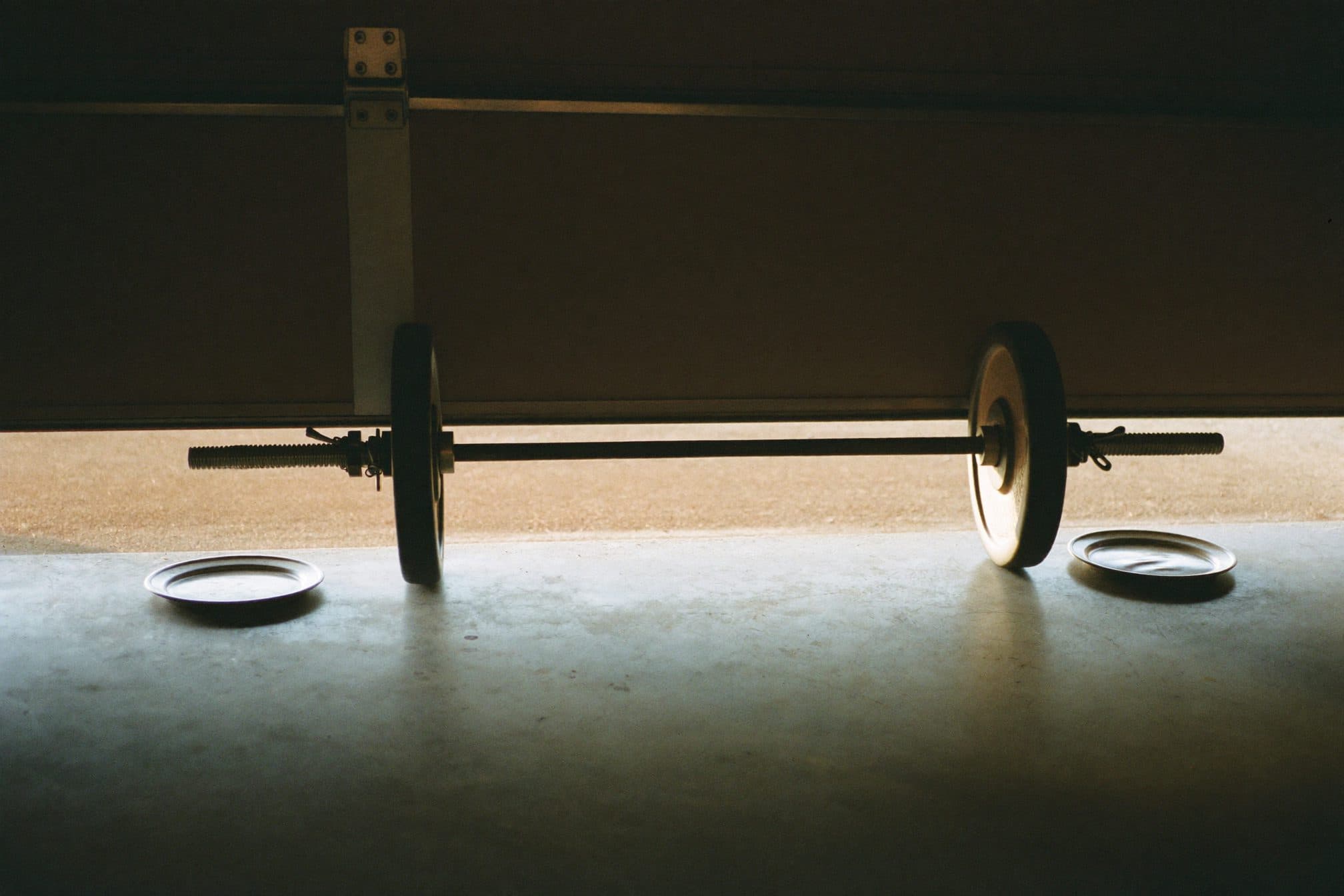 A barbell loaded with one modest plate per side on a garage floor, morning light under the door