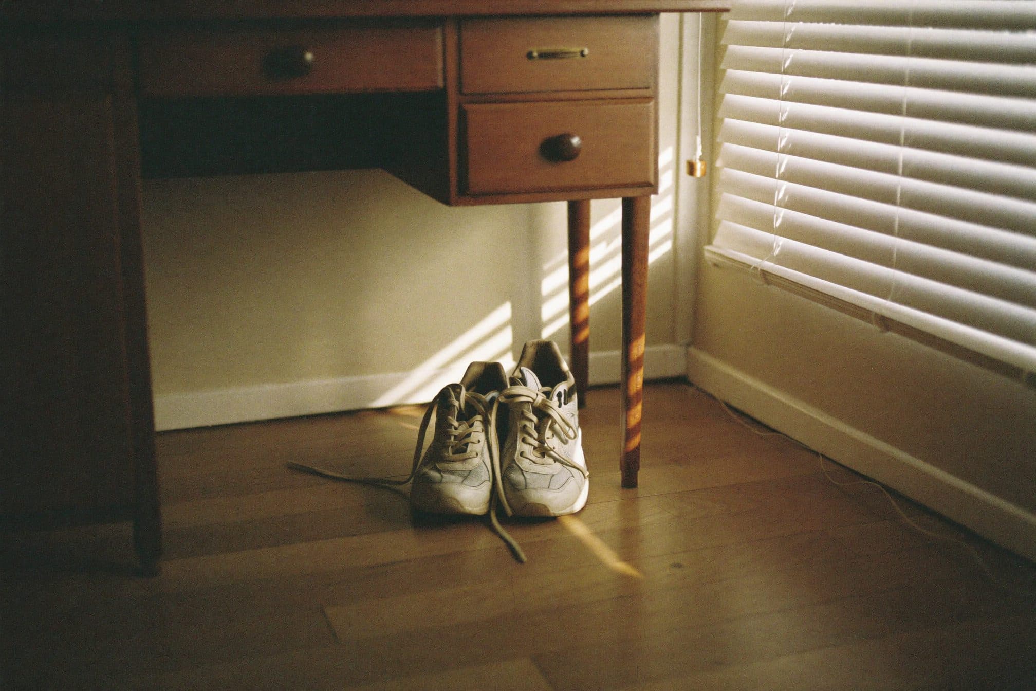 Worn sneakers tucked at the base of a desk, morning light striping the floor through blinds
