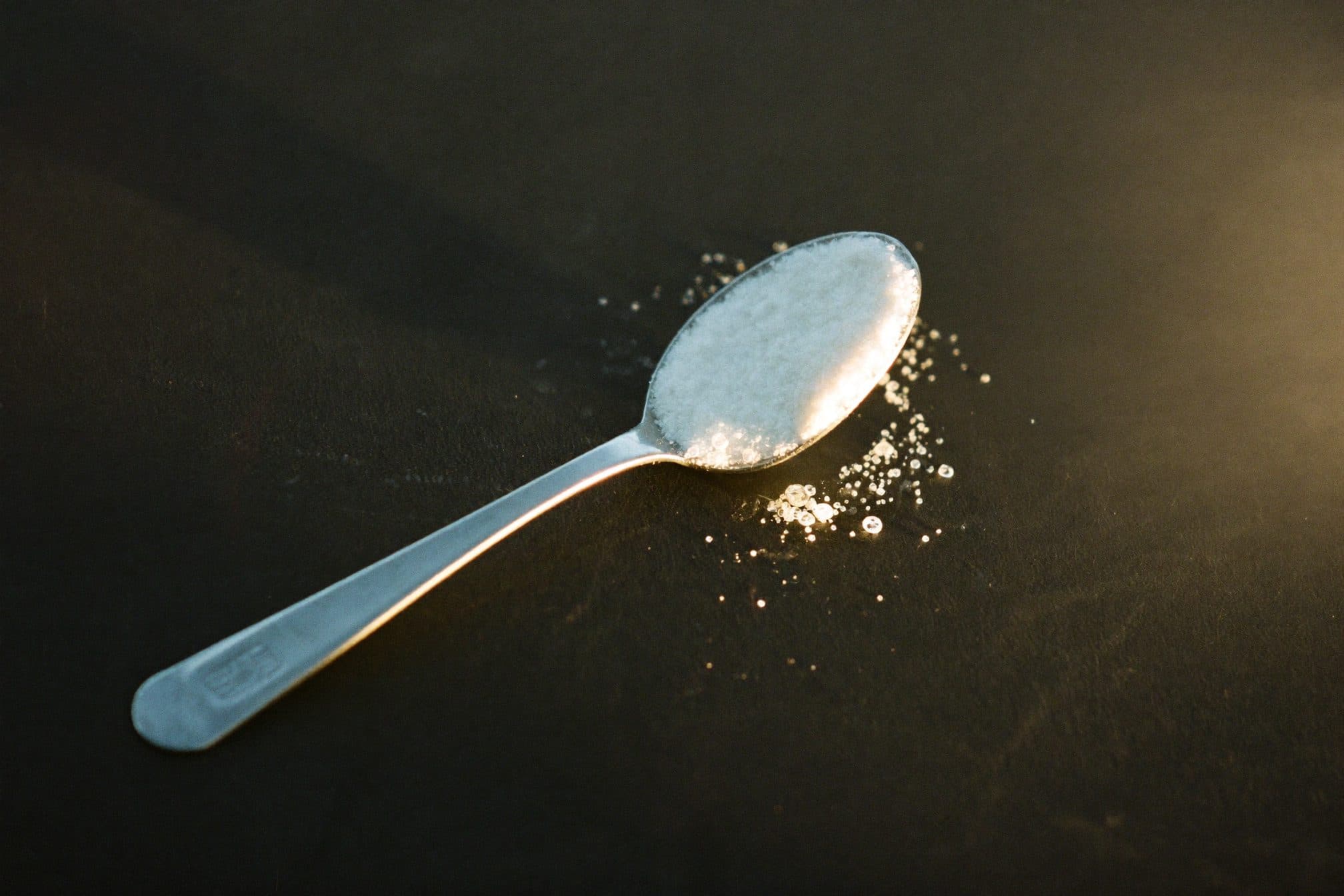 A level stainless teaspoon of fine white crystalline powder on dark slate in warm side light