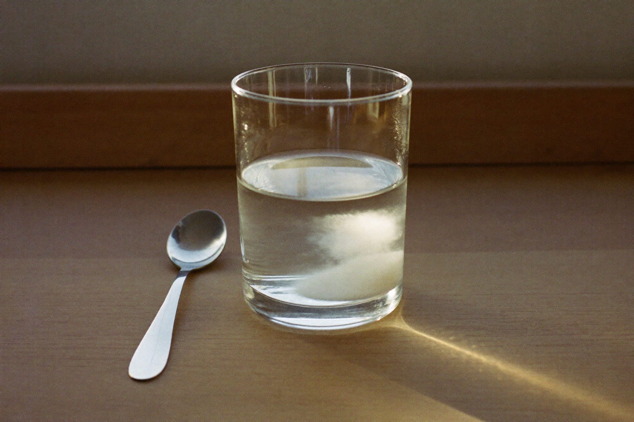 A glass of water with a faint swirl of dissolving white powder, spoon resting beside it