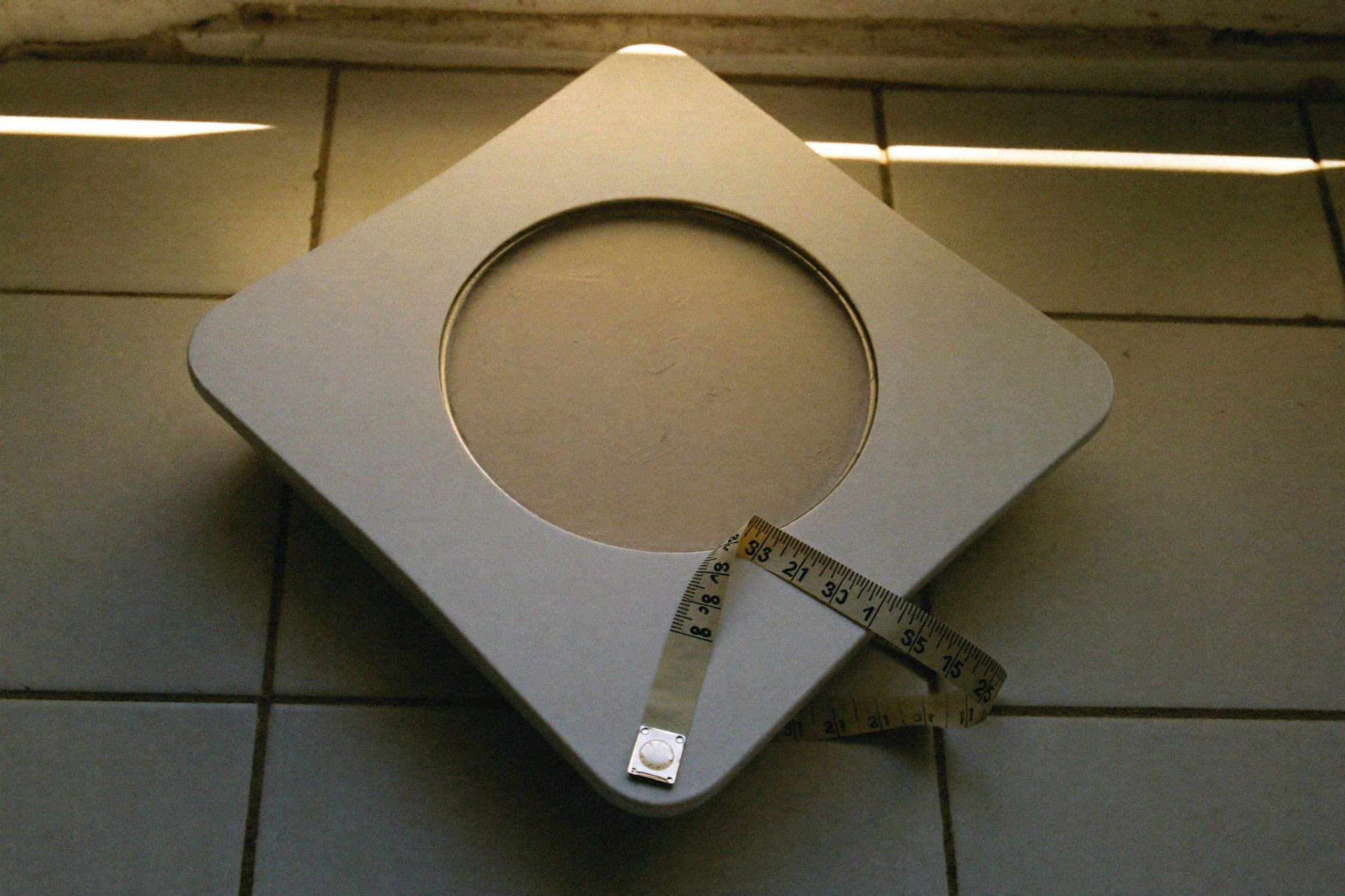A plain bathroom scale on ceramic tile with a cloth tape measure draped across one corner