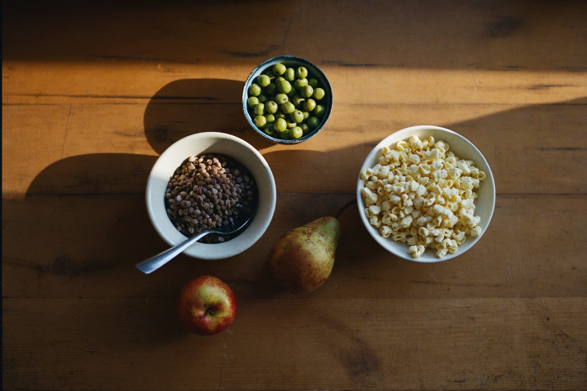 Overhead row of lentils, peas, popcorn, an apple, and a pear on worn wood in warm morning light