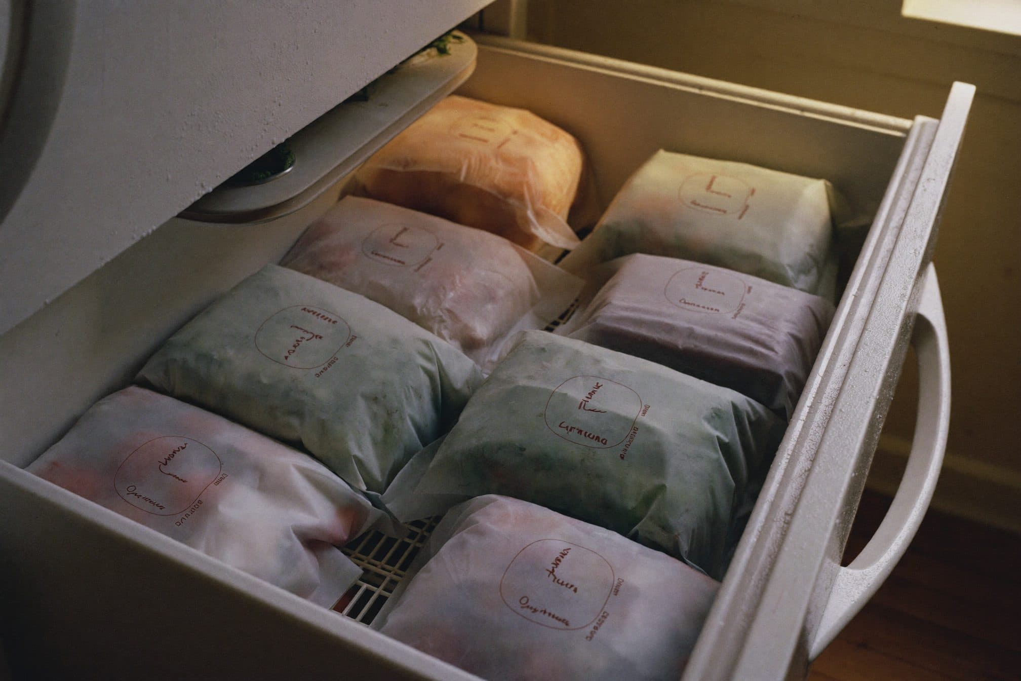 A home freezer drawer open with plain frosted bags of vegetables inside, cold glow meeting warm kitchen light