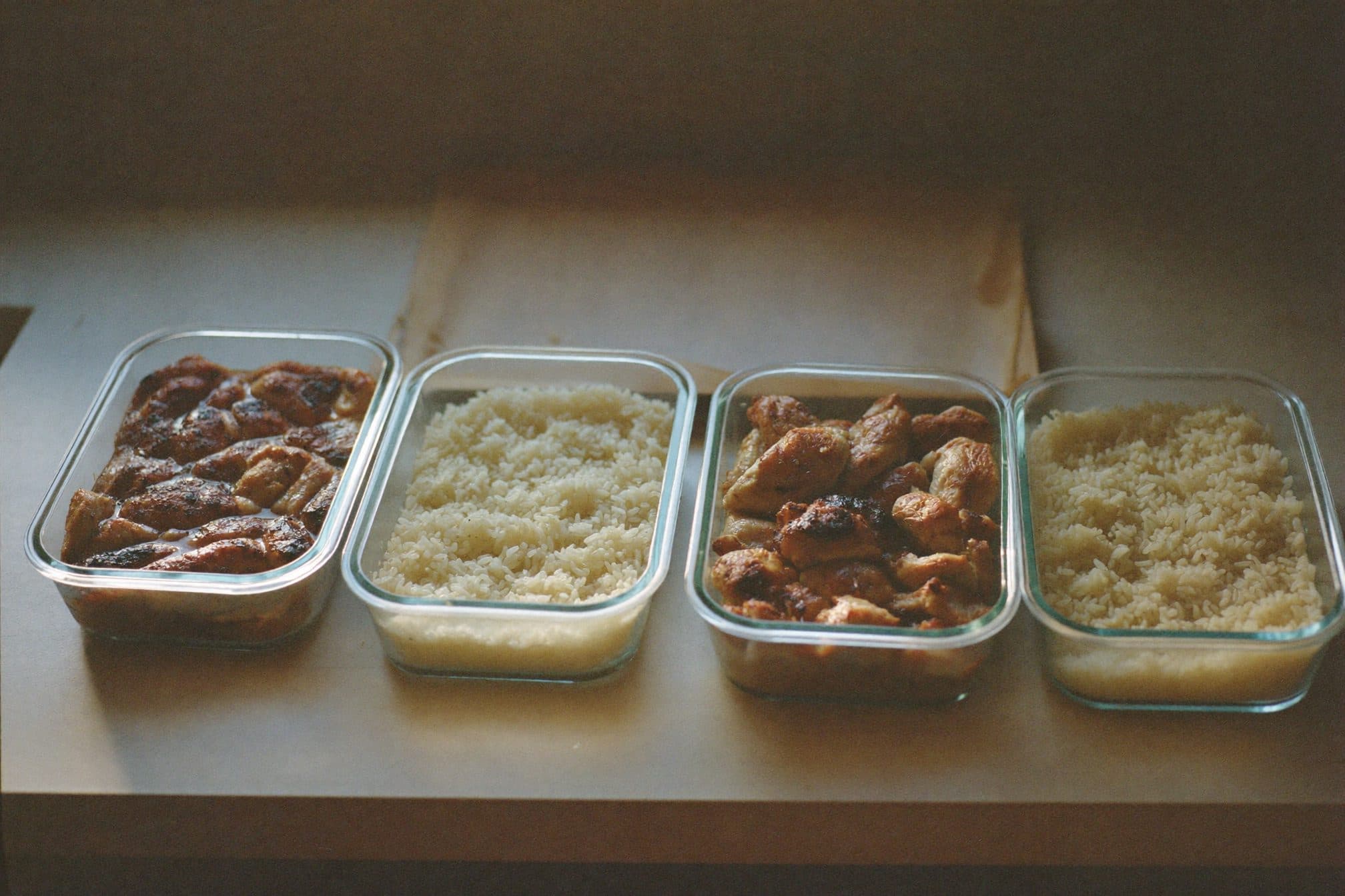 Four glass meal-prep containers of chicken and rice in a row, sheet pan behind, morning window light