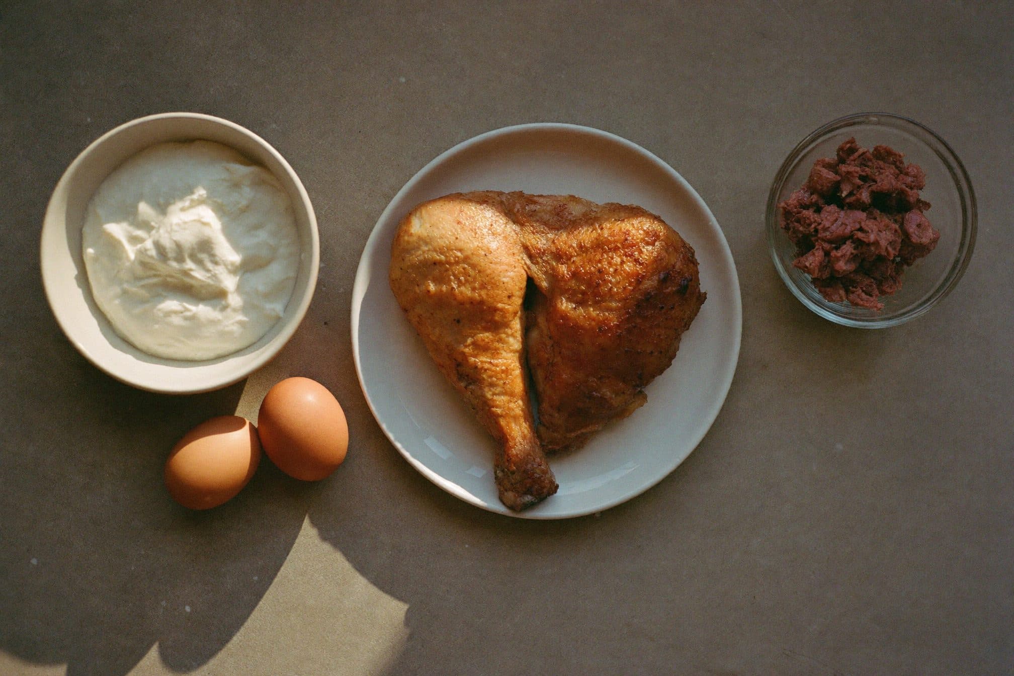 Overhead still life of a chicken thigh on a plate, a bowl of yogurt, two eggs, and a small bowl of tuna on a stone counter