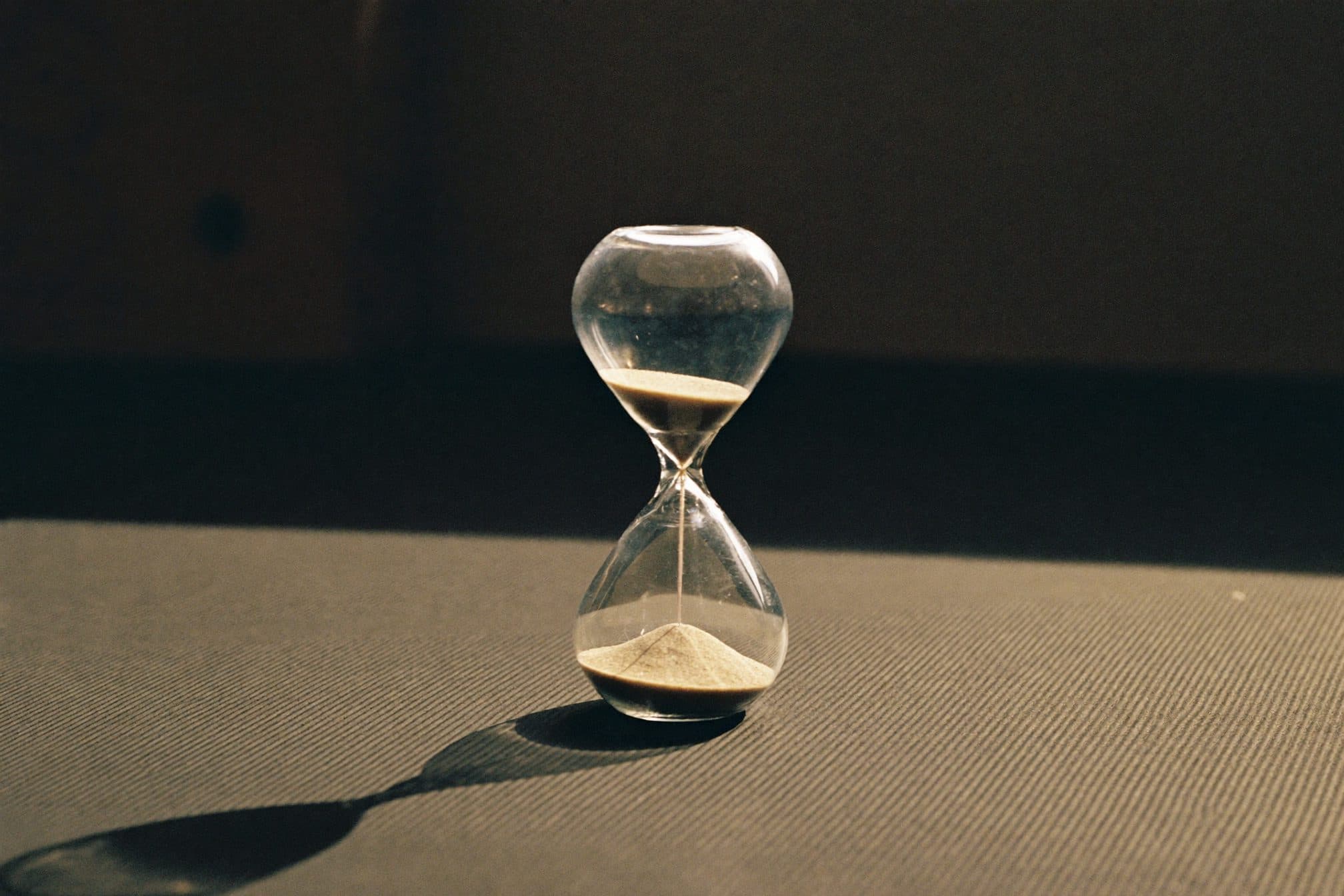 A small hourglass with pale sand half-run beside a black hex dumbbell on a rubber mat in warm morning light