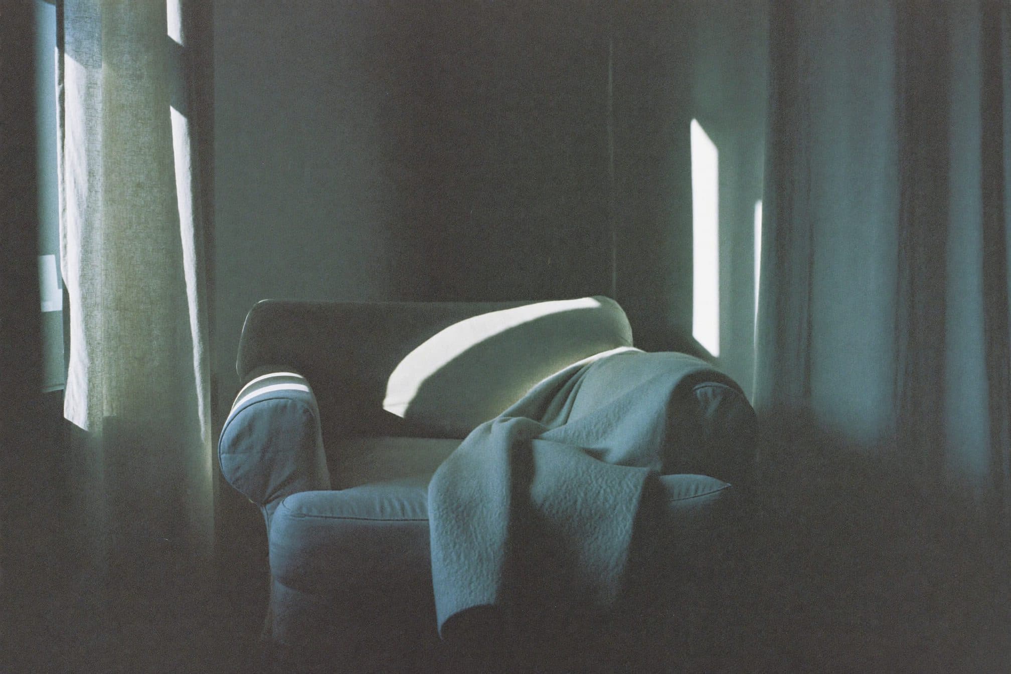 A dim living room with curtains nearly drawn, one blade of light across an empty armchair and folded blanket