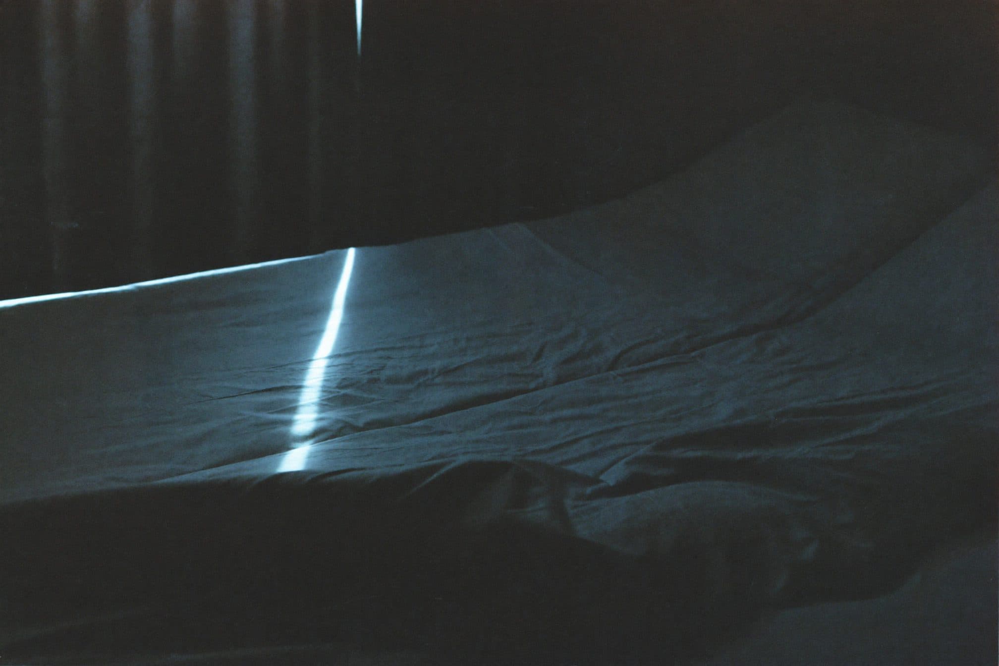 A rumpled empty bed at grey dawn, one seam of cold blue light across creased linen