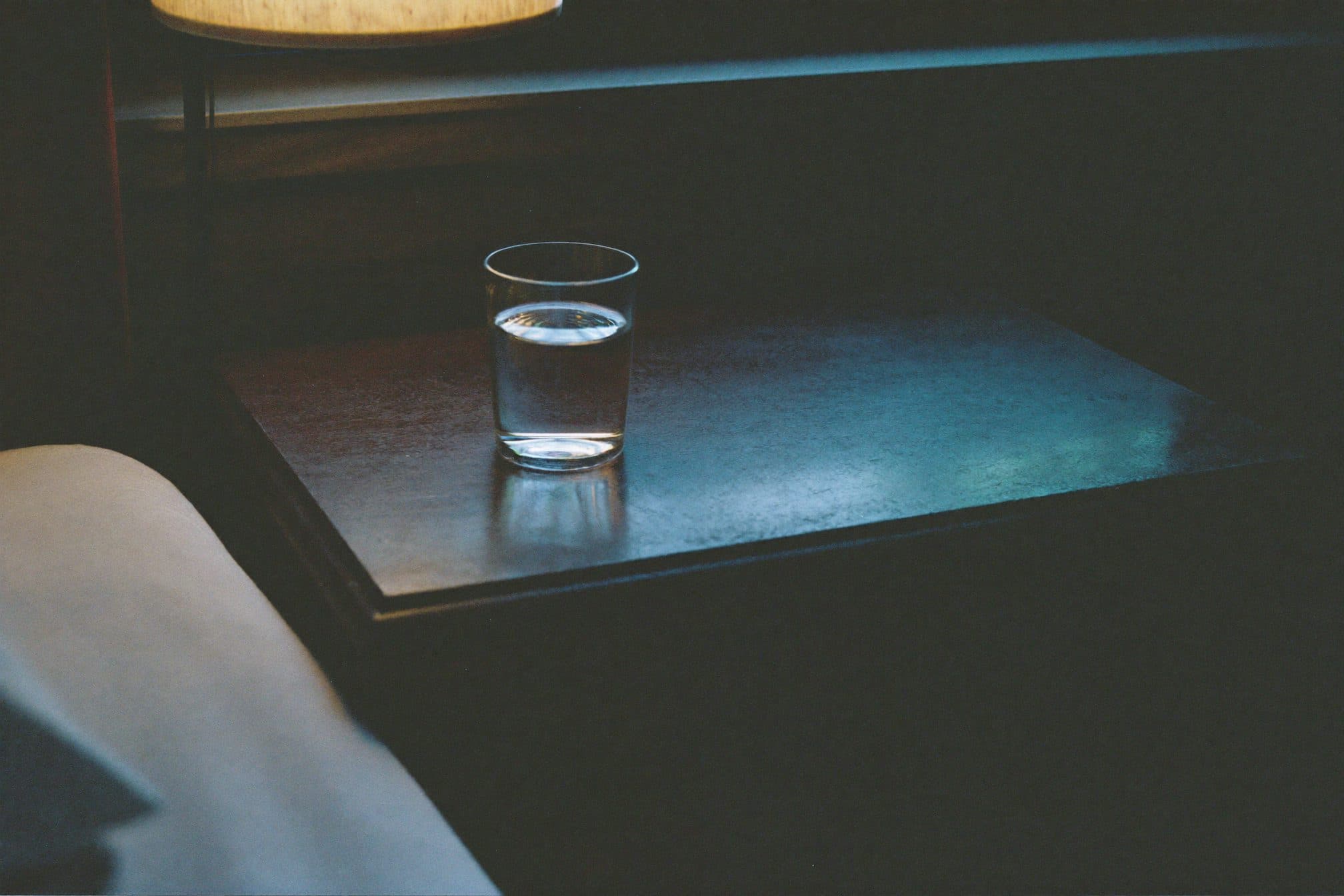 A single glass of water on a dark nightstand in dim blue night light with one warm lamp glow