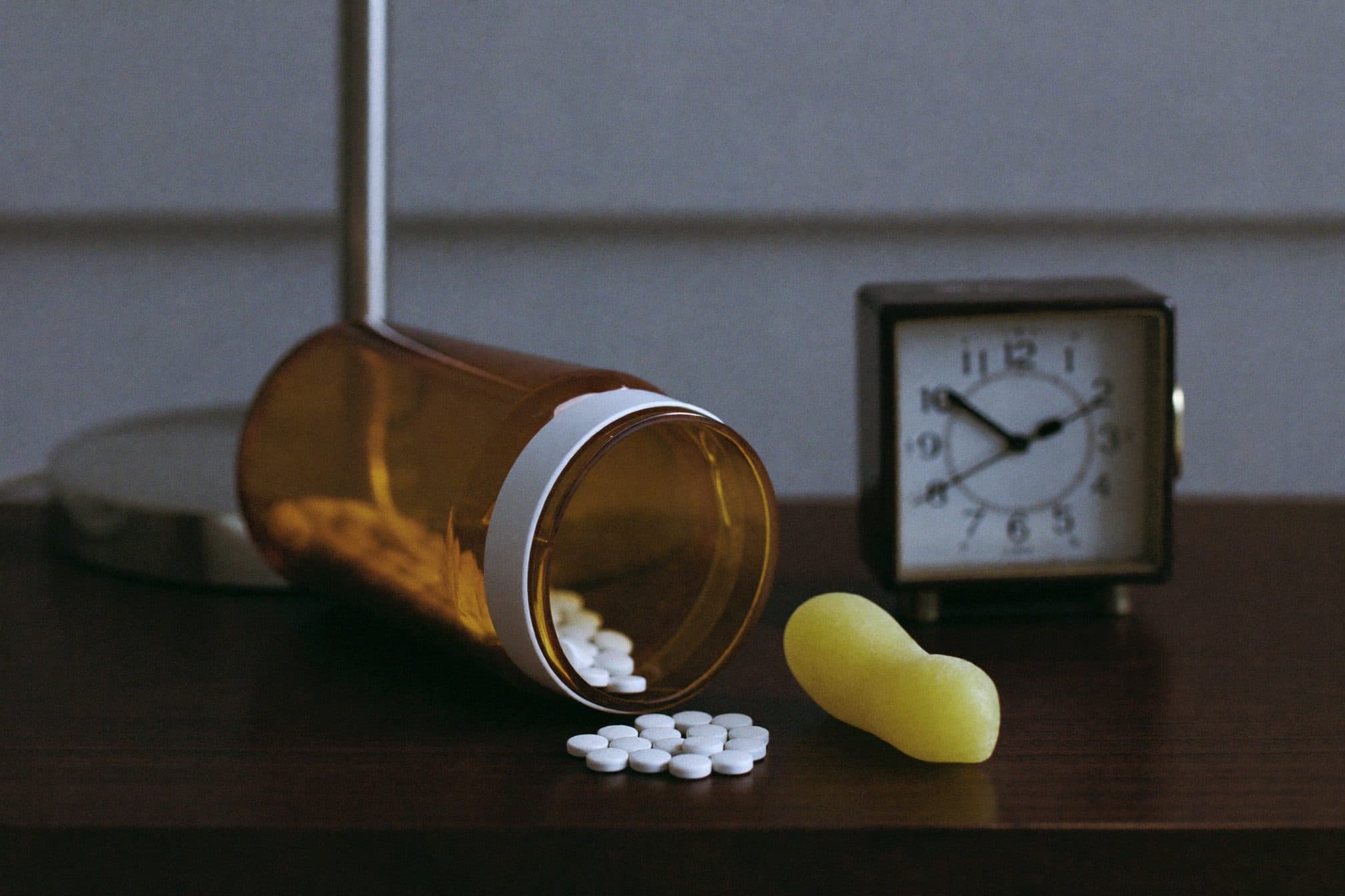 An amber pill bottle tipped on its side, tiny white tablets scattered beside one oversized gummy in cool lamp light