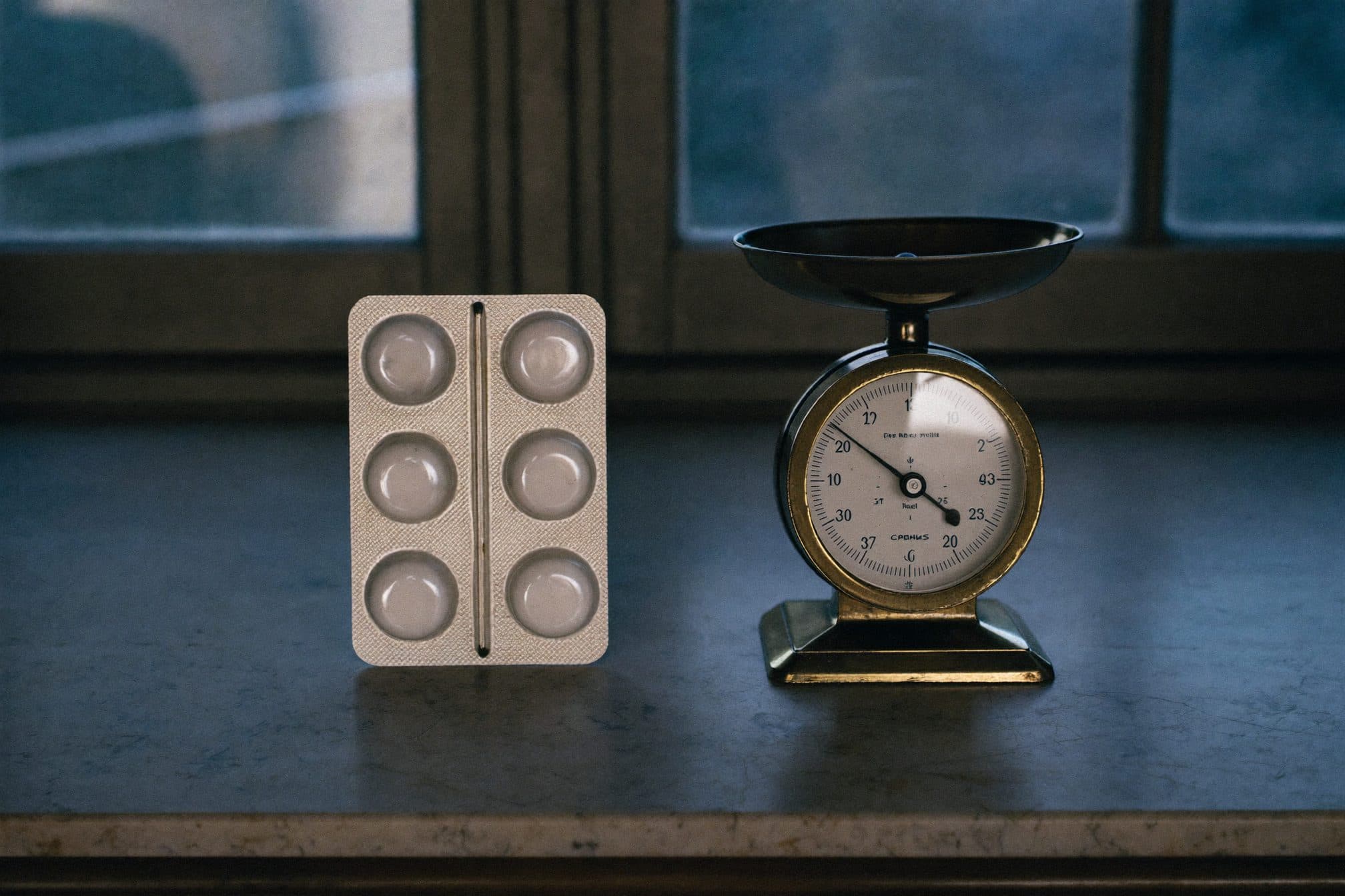 A foil blister pack of small tablets beside a small brass balance scale under blue-hour window light