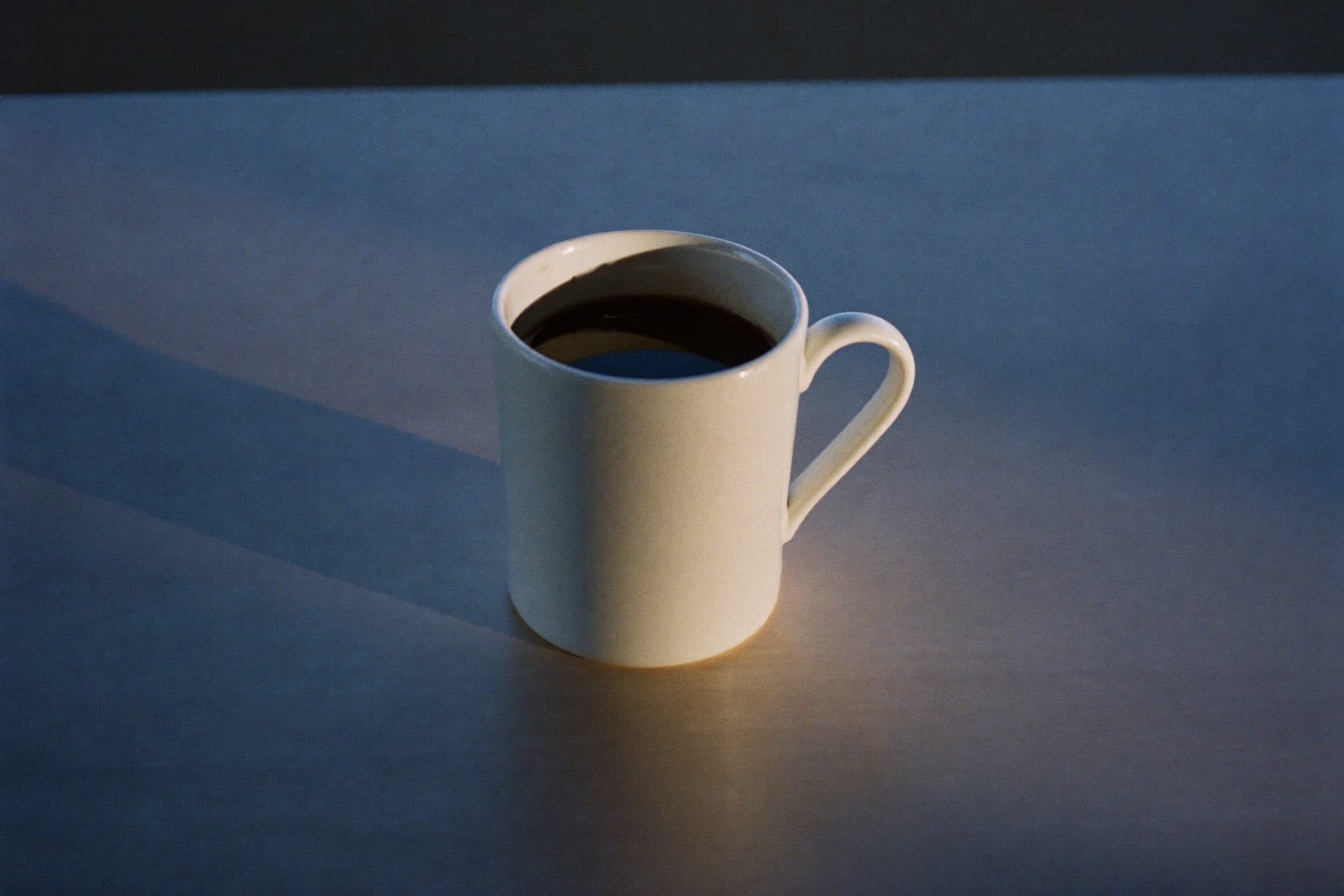 A plain white mug of coffee gone cold on a bare desk as afternoon light turns blue
