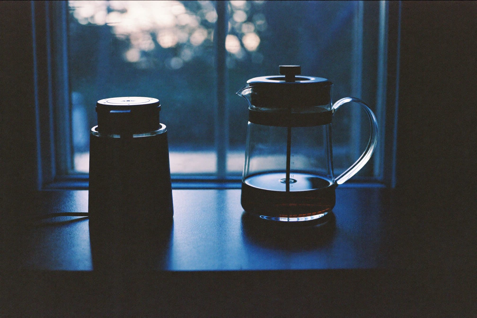A glass french press and unplugged grinder pushed to the back of a dark counter, moonlit window behind