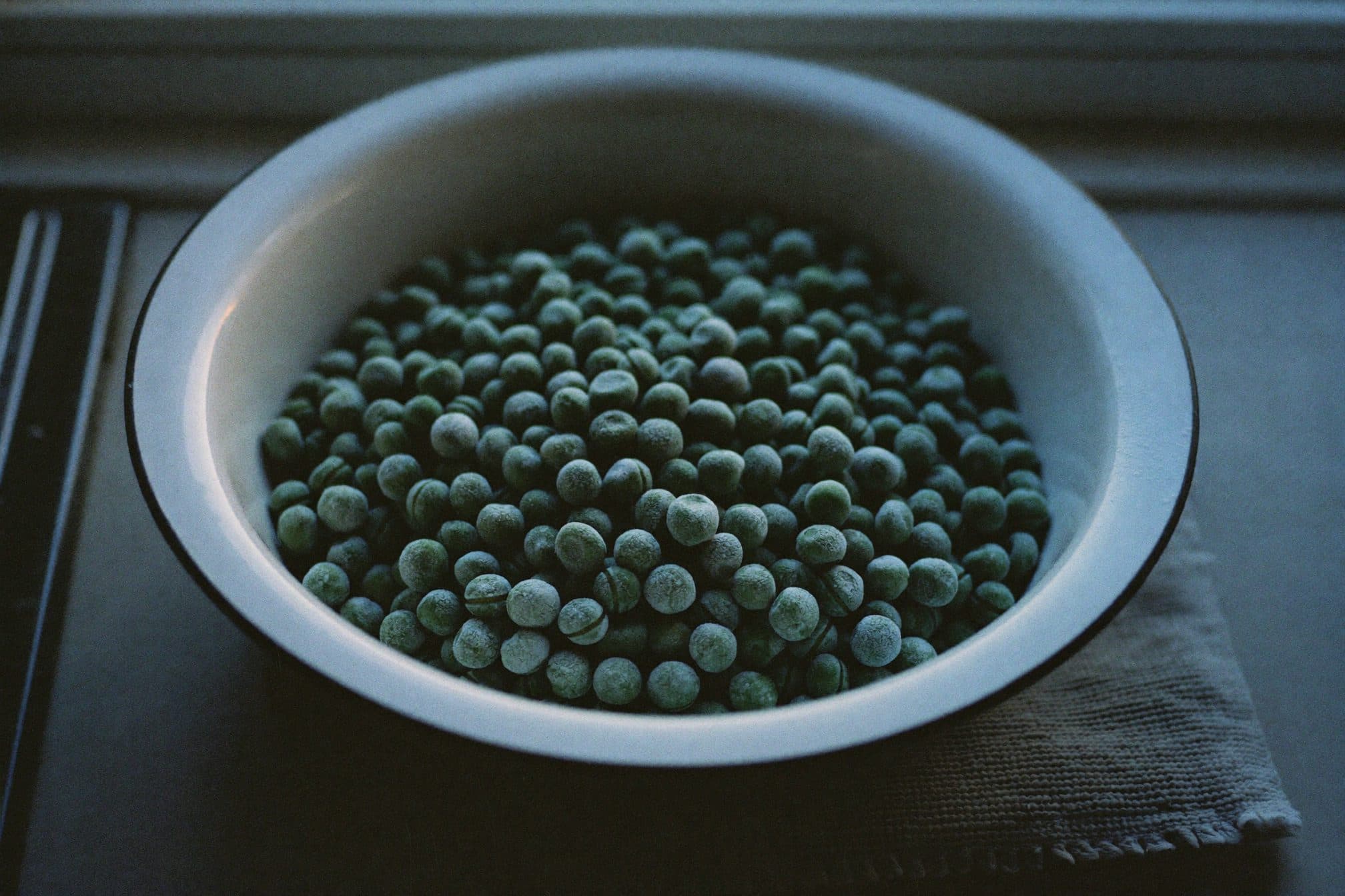 A white enamel bowl of frozen peas with frost haze on a folded kitchen towel in cool evening light