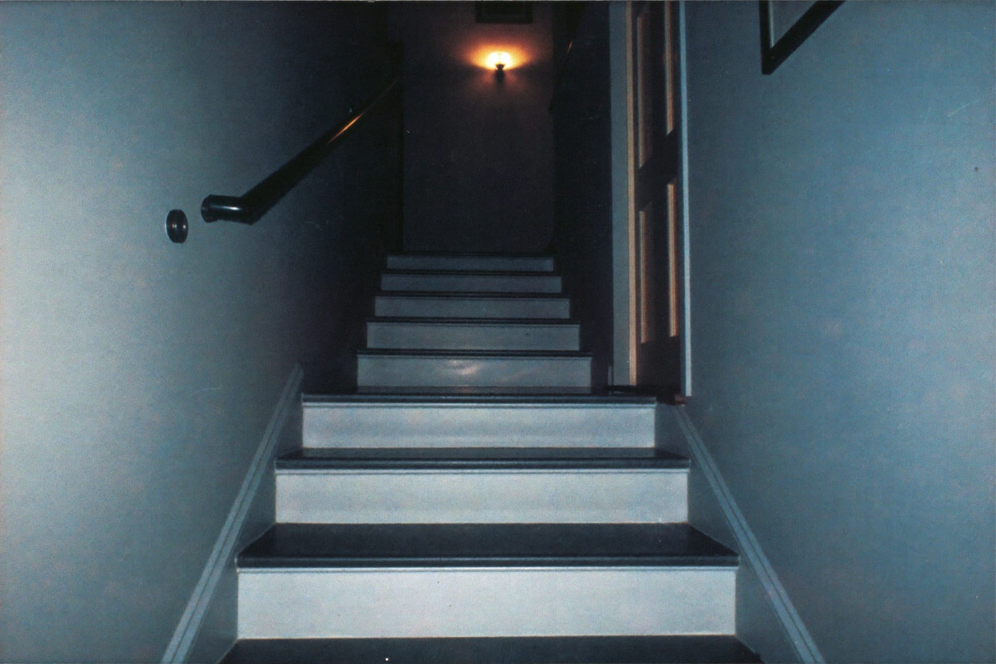 A home staircase at night in cool blue shadow, one small warm wall light halfway up