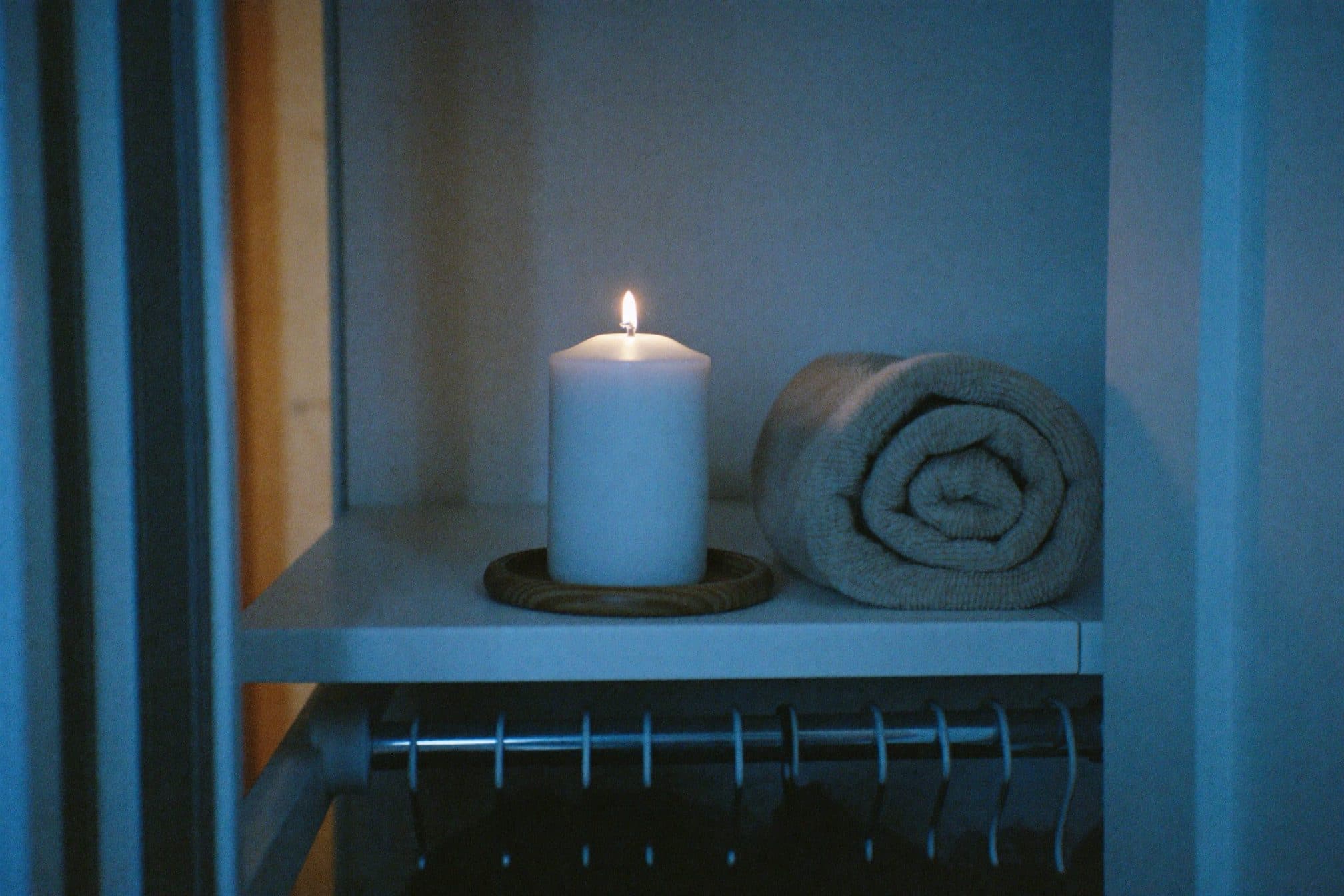 An unlit white candle on a wooden dish beside a rolled grey towel on a shelf in cool evening light