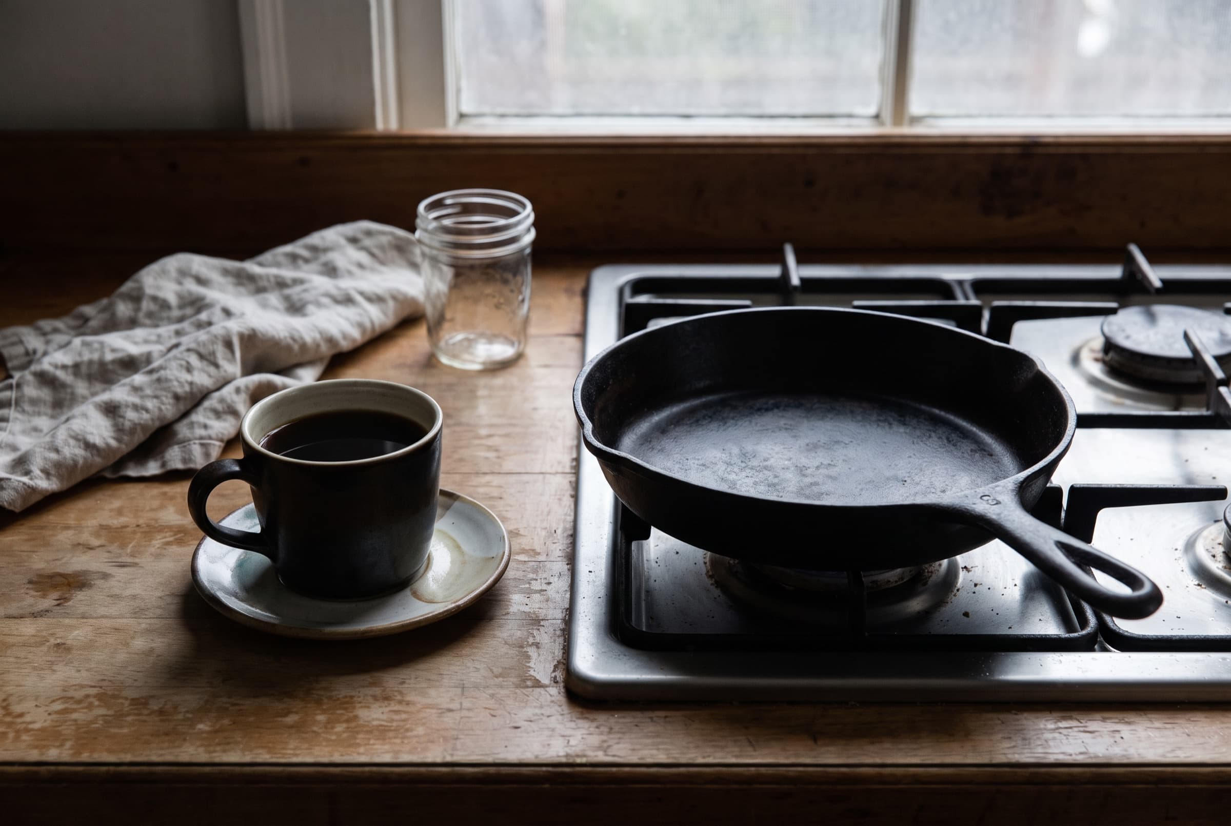 A black coffee beside a cold cast-iron pan on an unlit stove in quiet grey morning light