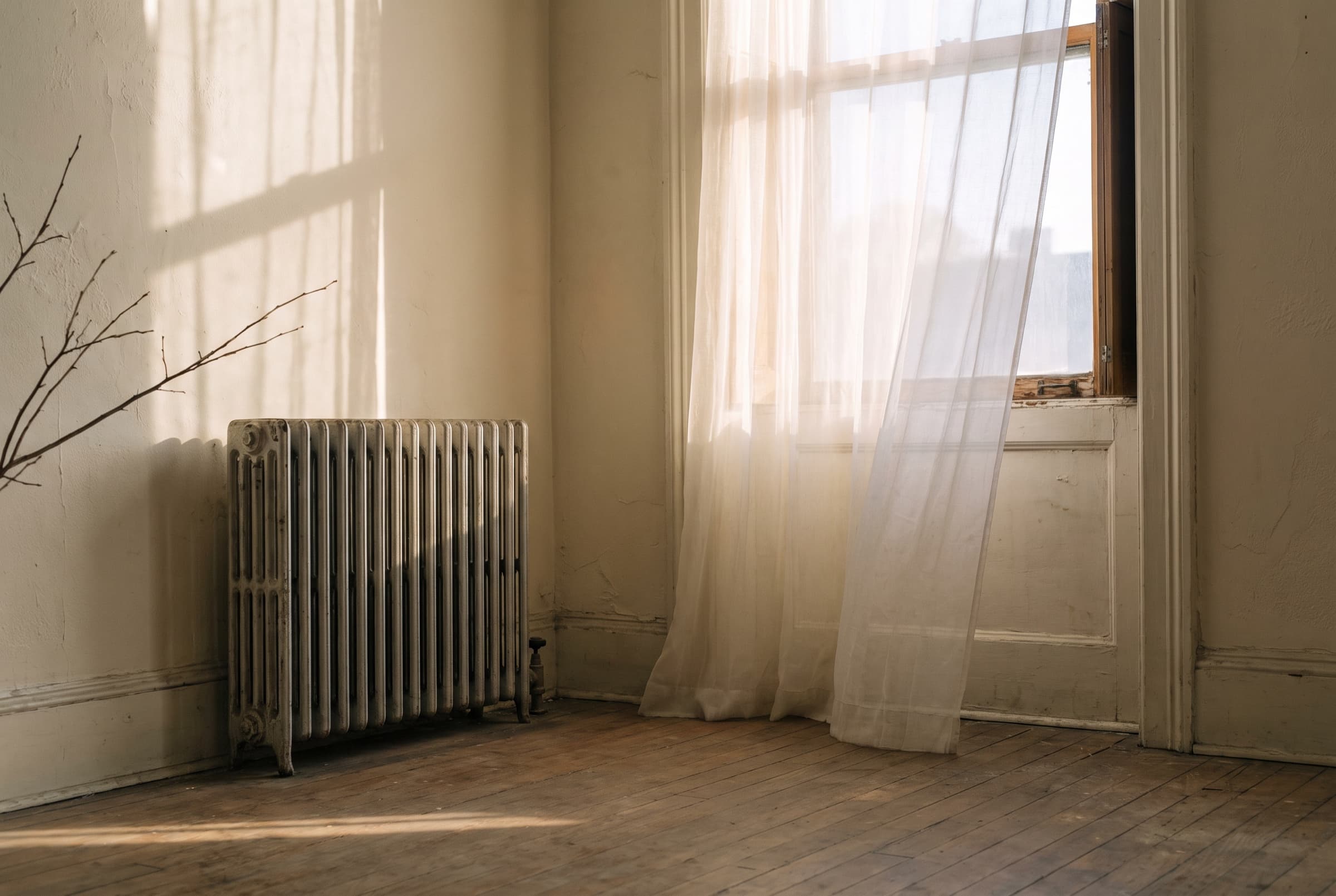 A sheer curtain glowing with low winter sun above an old radiator in a quiet room, long shadows on the floor