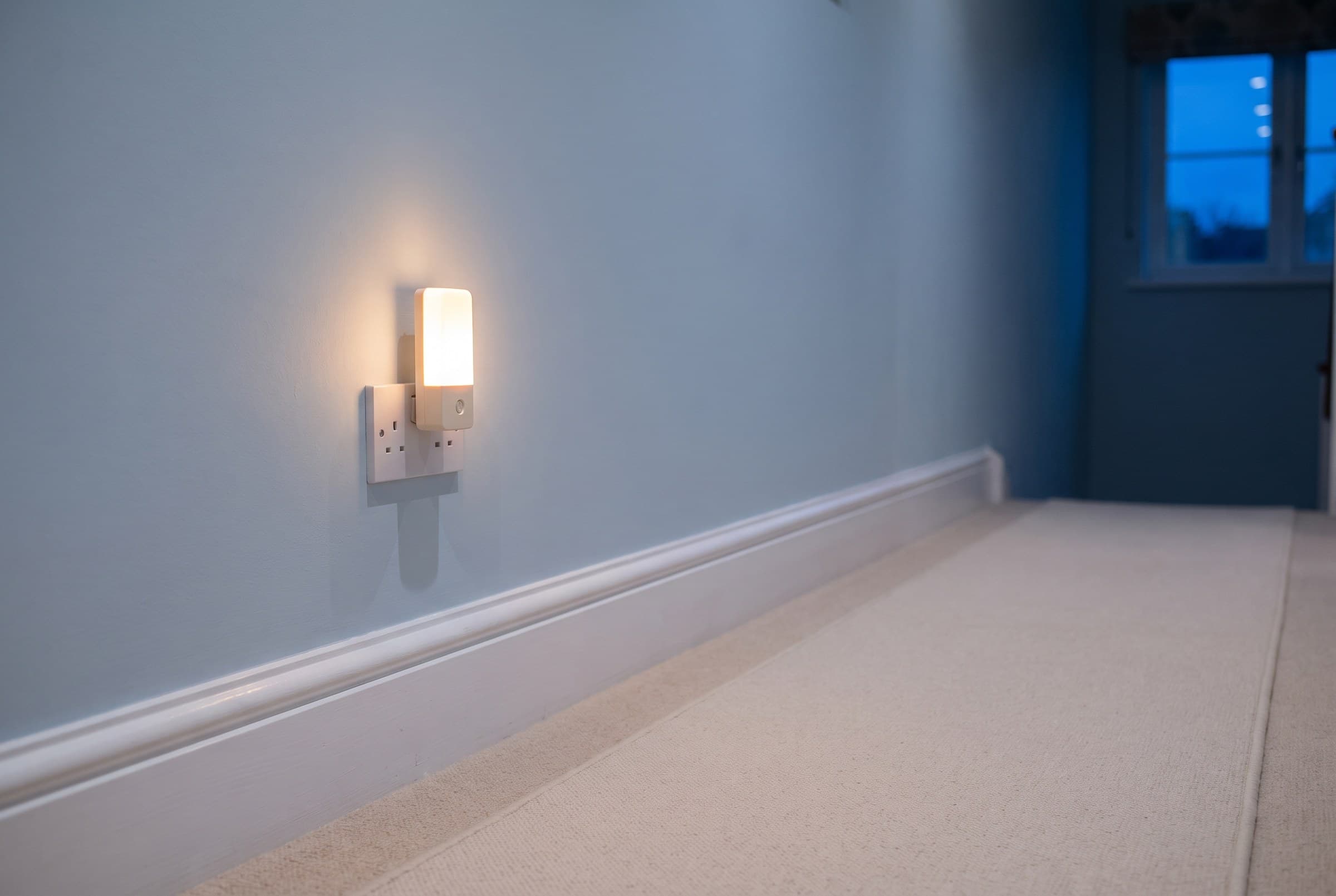 A small plug-in nightlight glowing warm on a pale hallway wall above a white skirting board, clean carpet running away toward a deep blue window