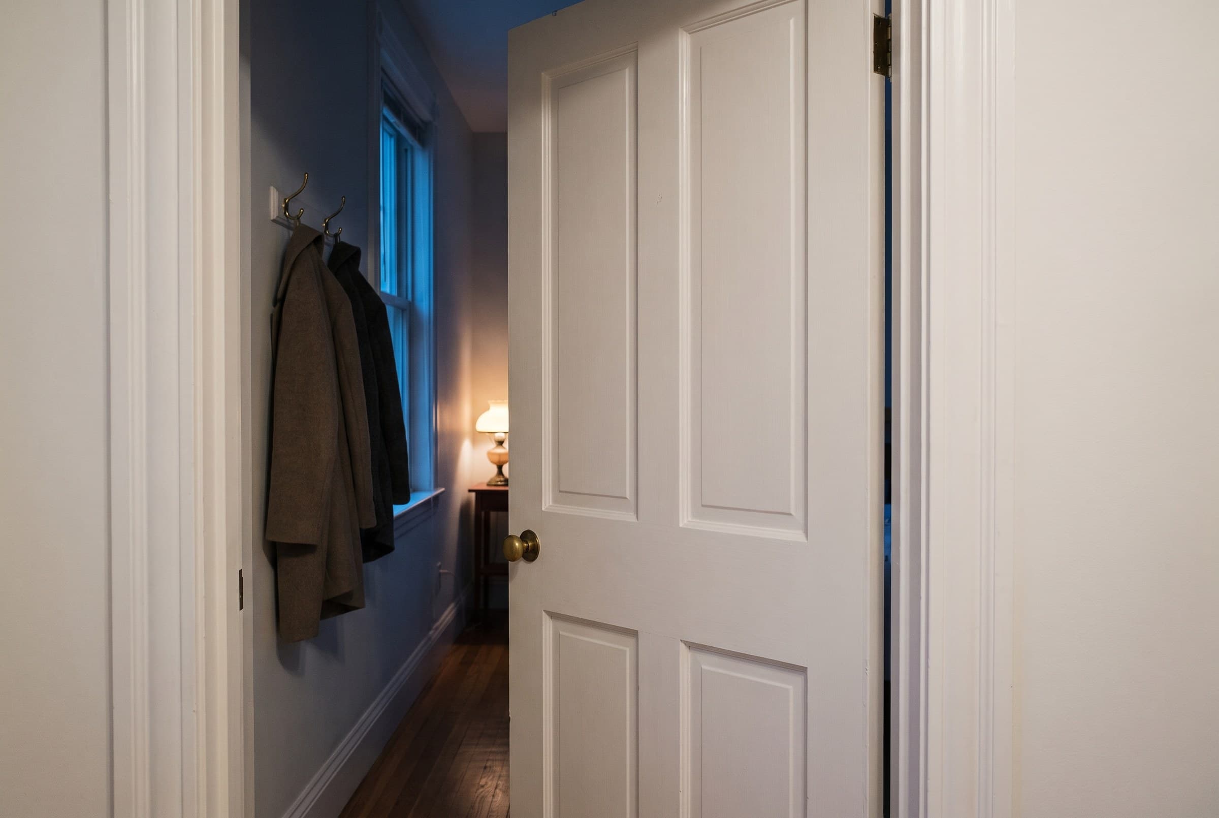 A white panel door standing ajar on a brass knob, opening onto a dim hallway with coats hung on hooks and one warm lamp glowing at the far end