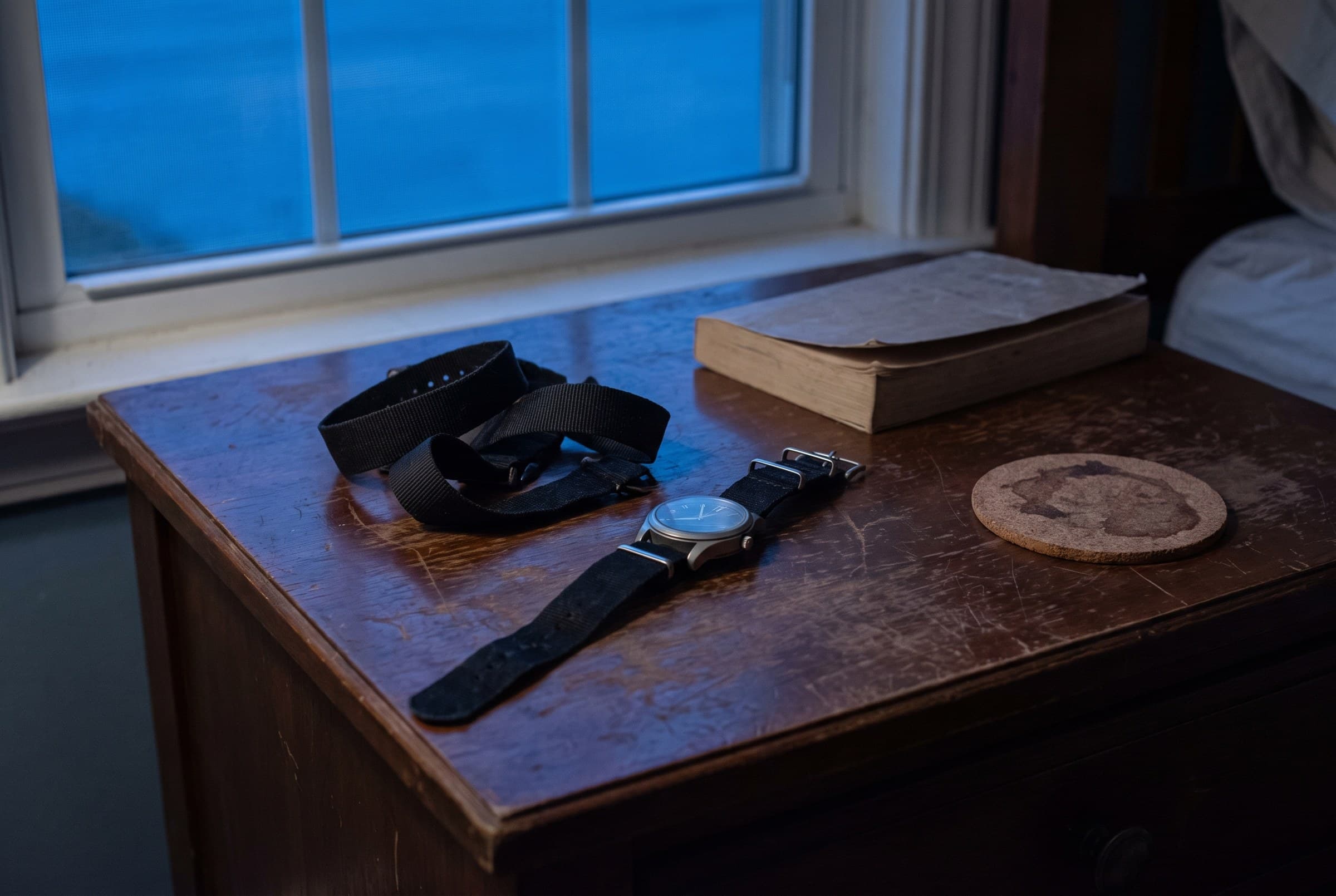 A plain watch with a dark unlit dial lying on a scratched wooden nightstand beside a slack fabric strap, a worn paperback and a stained coaster